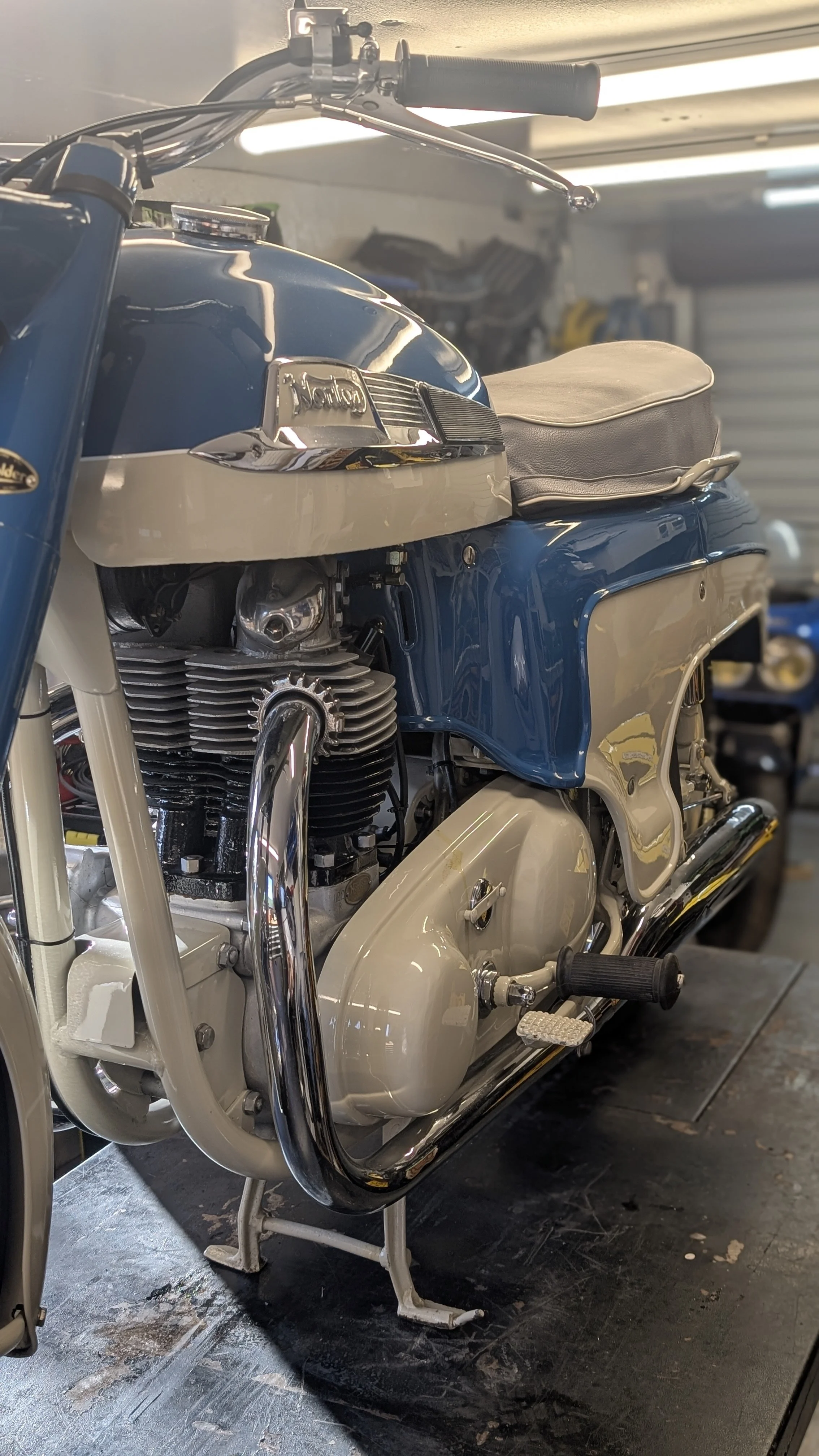 Close-up of a vintage motorcycle with a shiny blue and white paint job, chrome details, and a black seat, located in a garage.