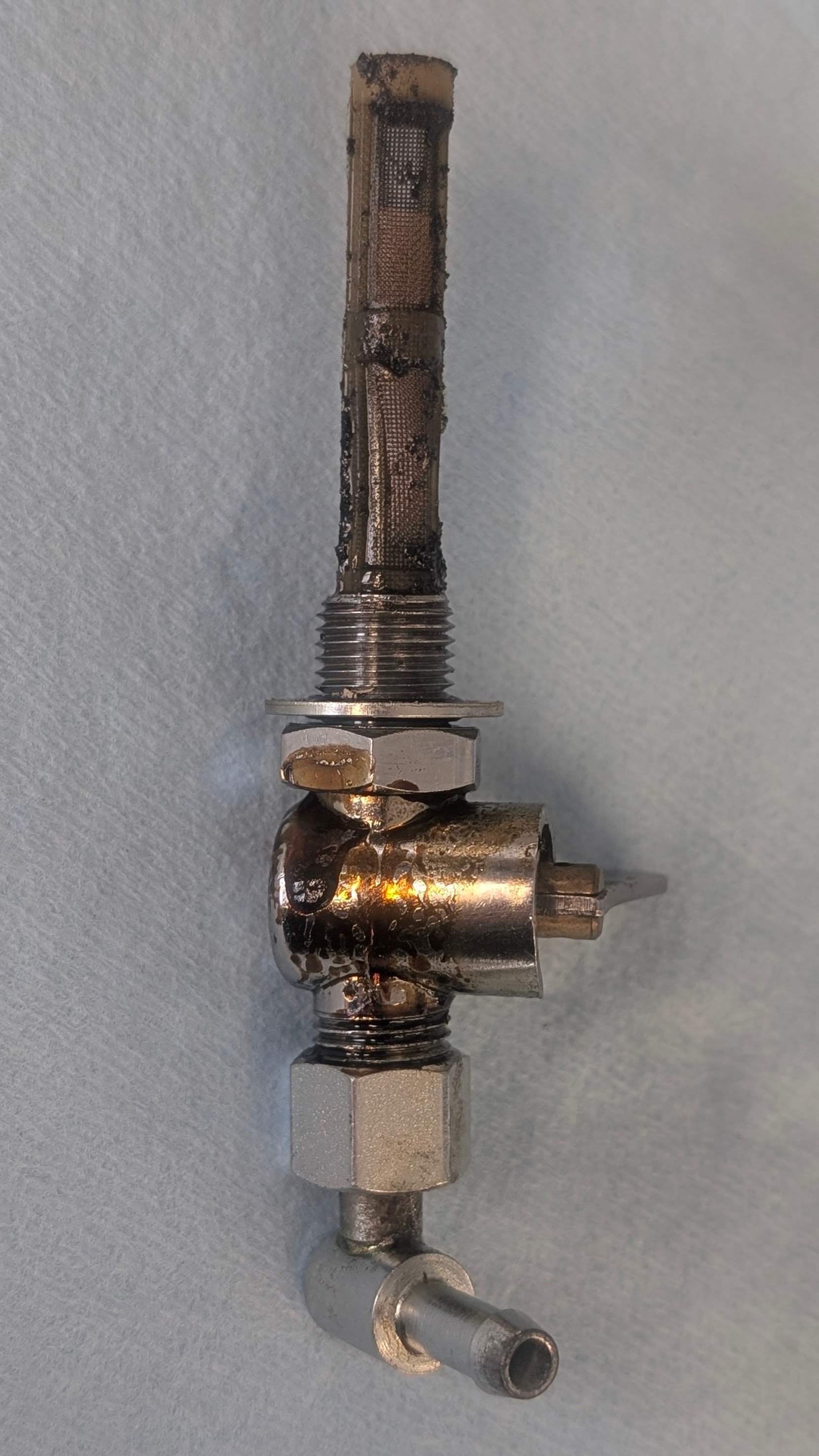 A metallic mechanical component with a threaded section, a curved metallic tube, and a hexagonal nut, covered in oil or grease, against a light background.