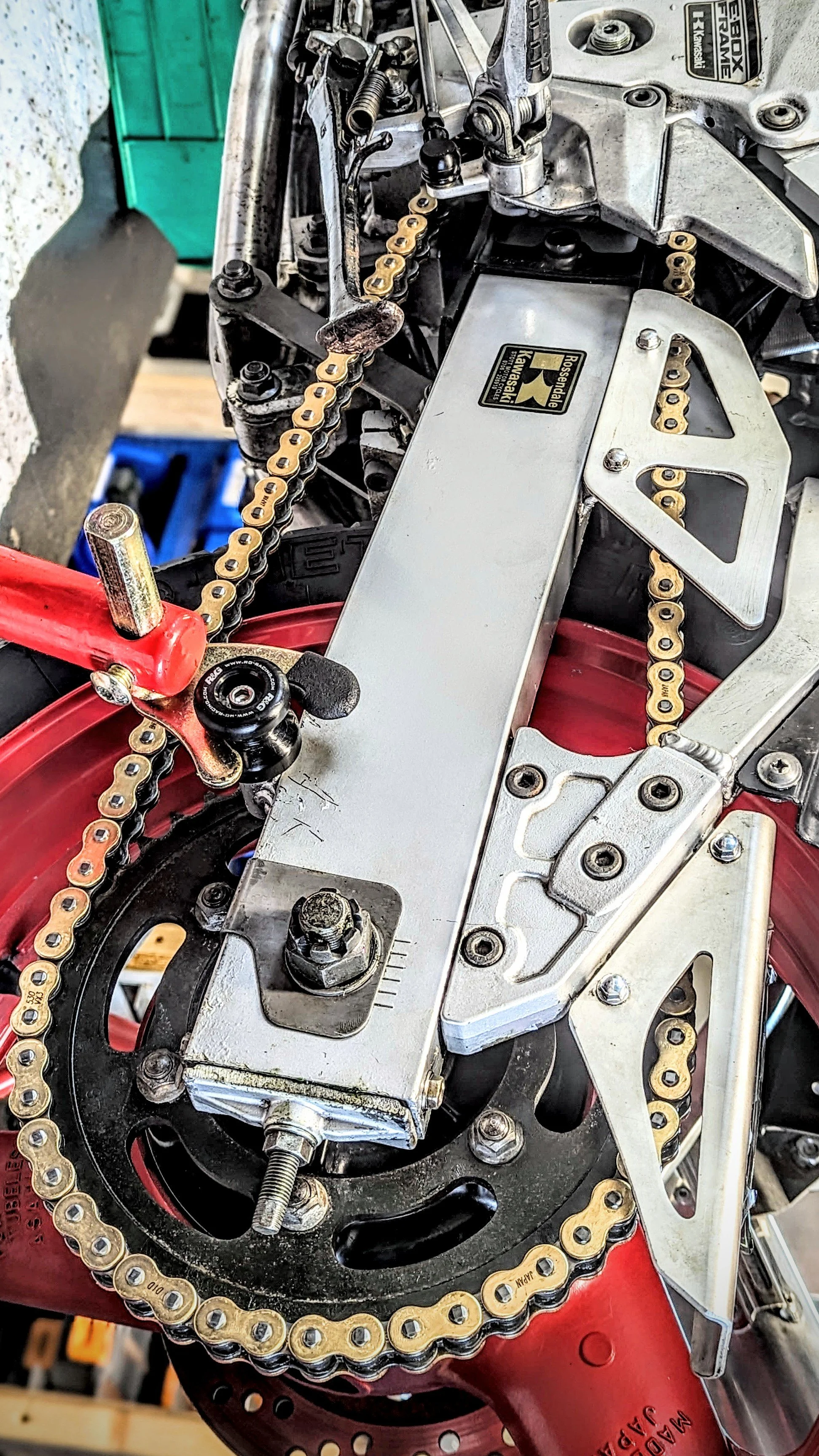 Close-up of a motorcycle chain, sprocket, and mechanical components with a metal link guard and various bolts and screws.