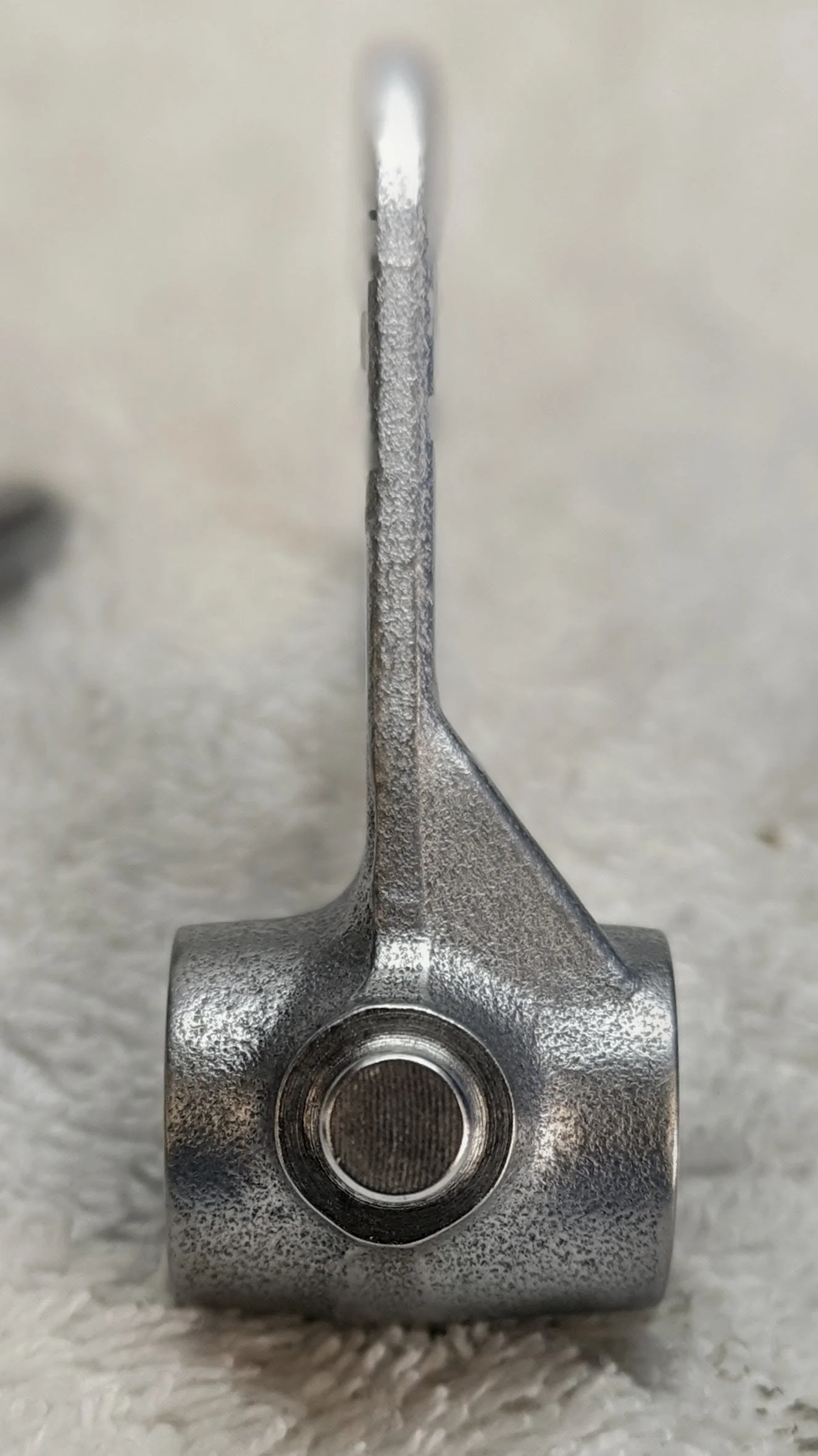 Close-up of a metallic mechanical component, possibly a valve or a part of a machine, with a cylindrical opening at the bottom and a long, narrow, textured stem extending upward, resting on a soft, white fabric surface.