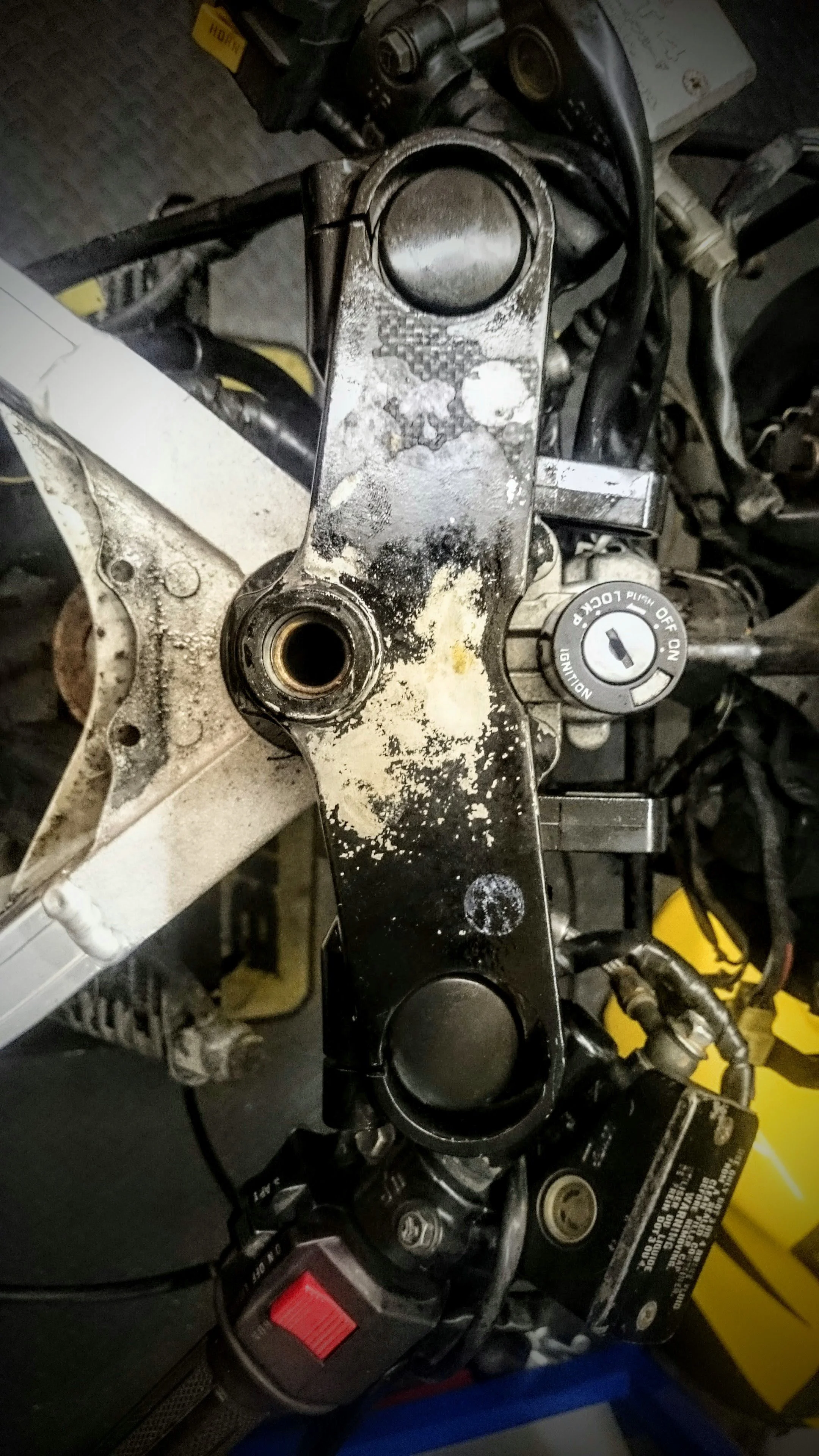 Close-up of a motorcycle's top triple clamp, showing a worn and dirty surface with missing handlebar grips, surrounded by various motorcycle components, wires, and tools in the background.