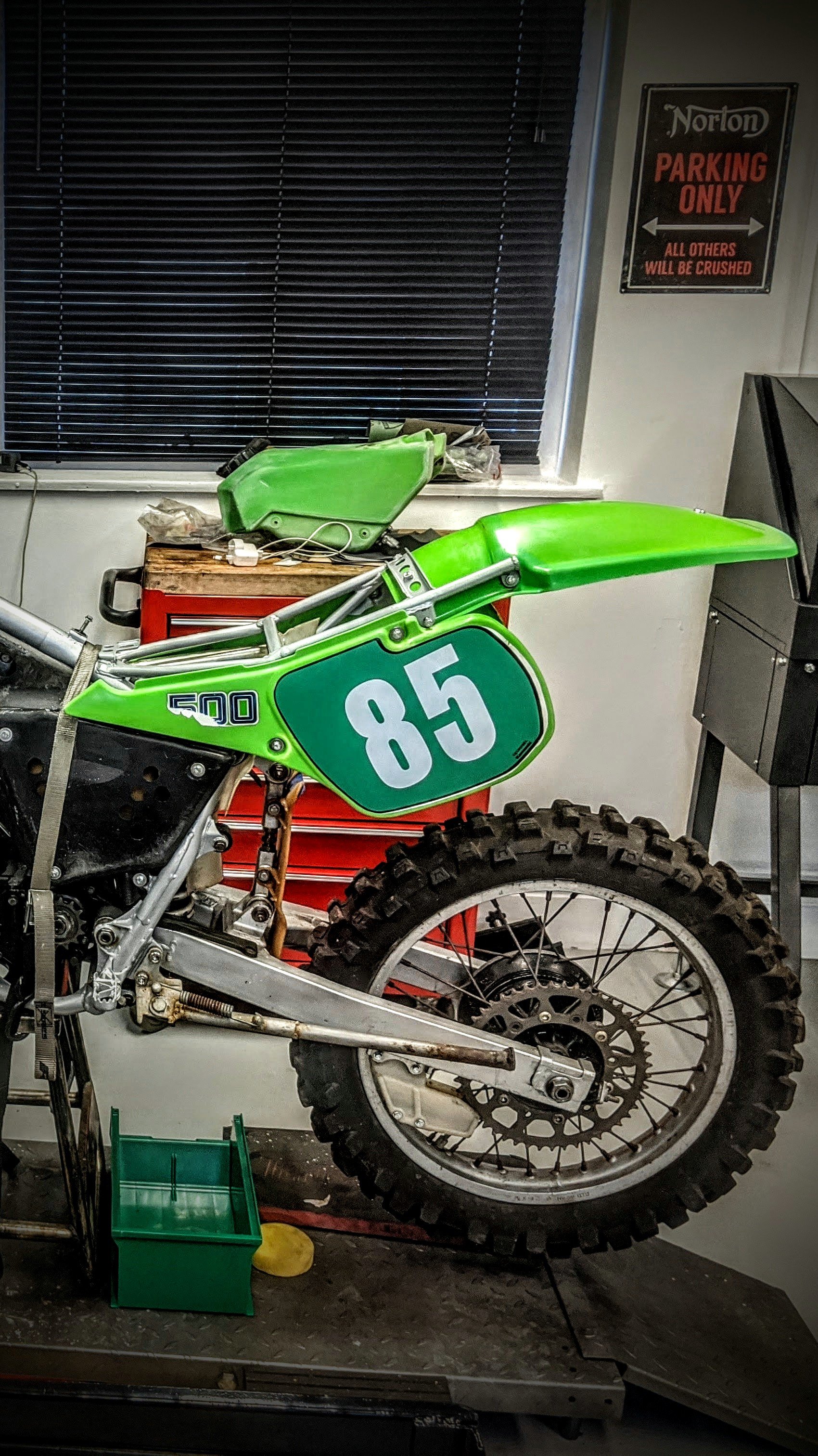 Green dirt bike displaying the number 85, situated in a workshop with a red tool chest, black window blinds, and a Nortlon parking only sign on the wall.