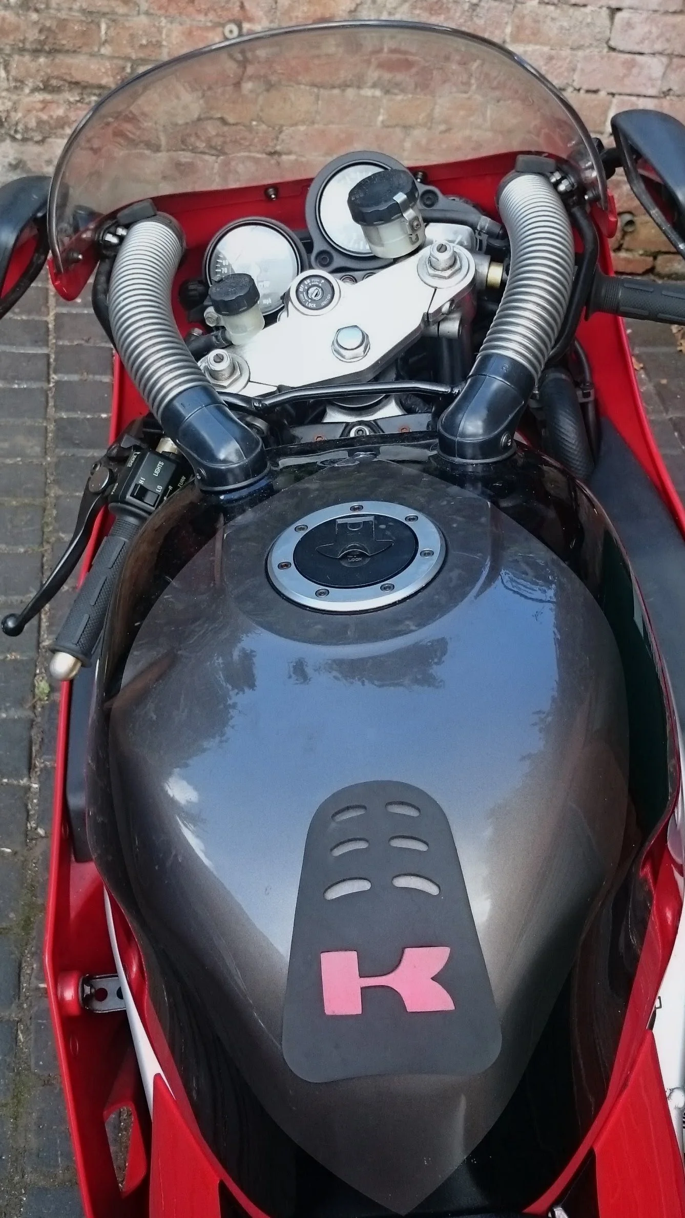 Top view of a motorcycle's dashboard, handlebars, and fuel tank with a black cover featuring a pink 'K' logo, all inside a red frame and a clear windshield, with a brick wall background.