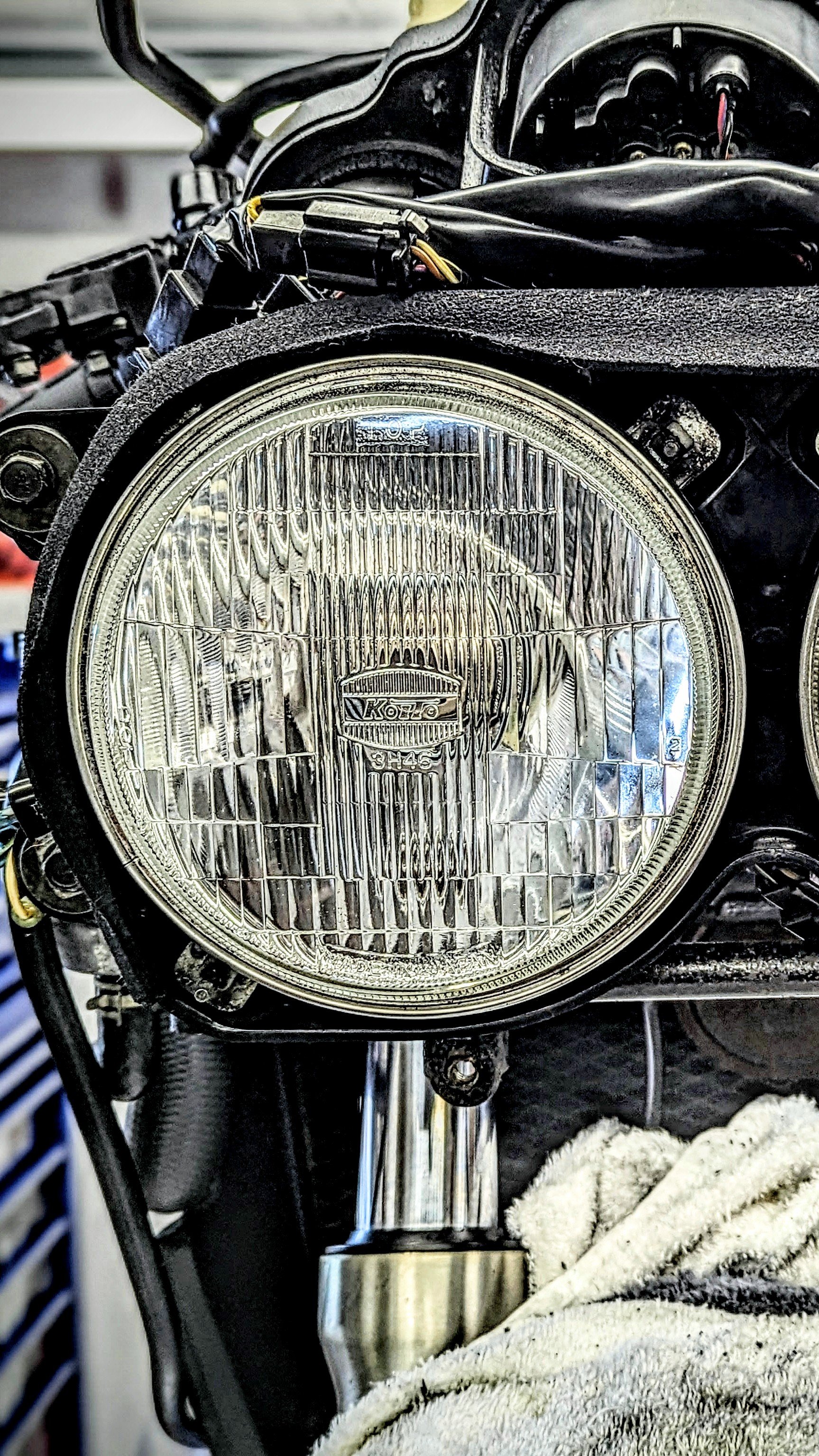 Close-up of a motorcycle headlight with surrounding wires and components, with a cloth underneath.
