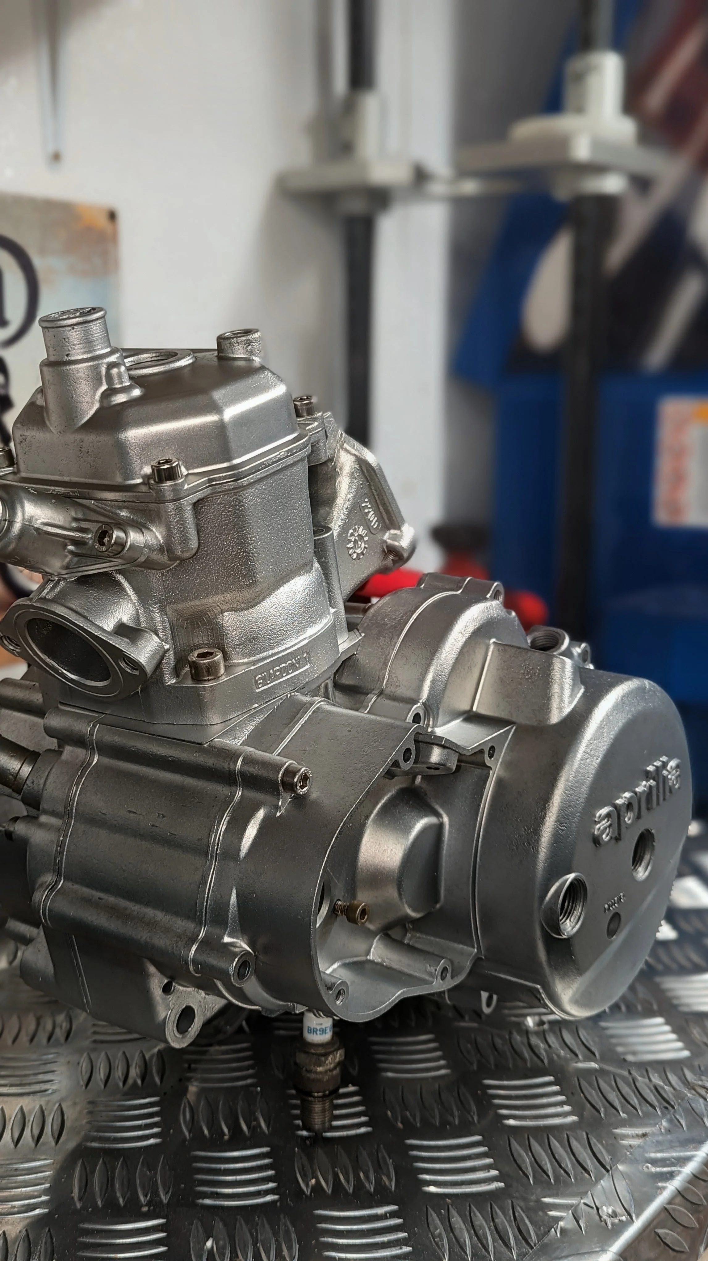 A close-up of a metallic engine or mechanical part placed on a textured surface, with blurred tools and equipment in the background.