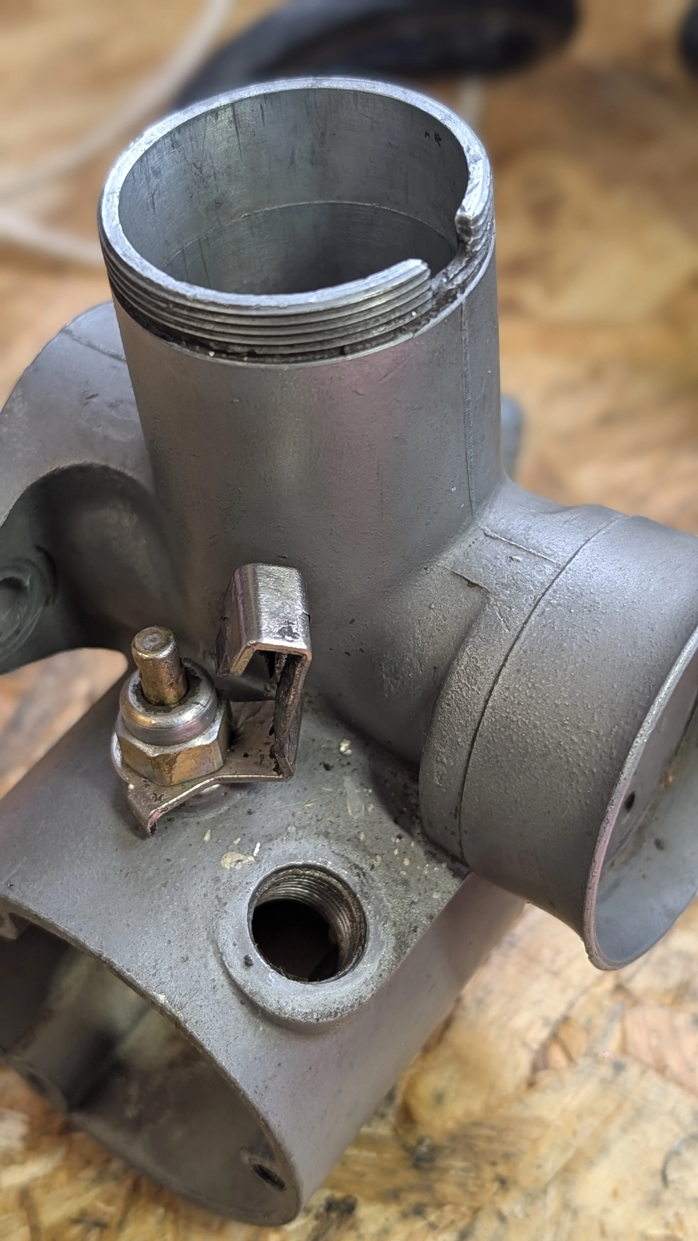 Close-up of a metallic carburetor with a visible opening and adjustment screw, placed on a wooden workbench.