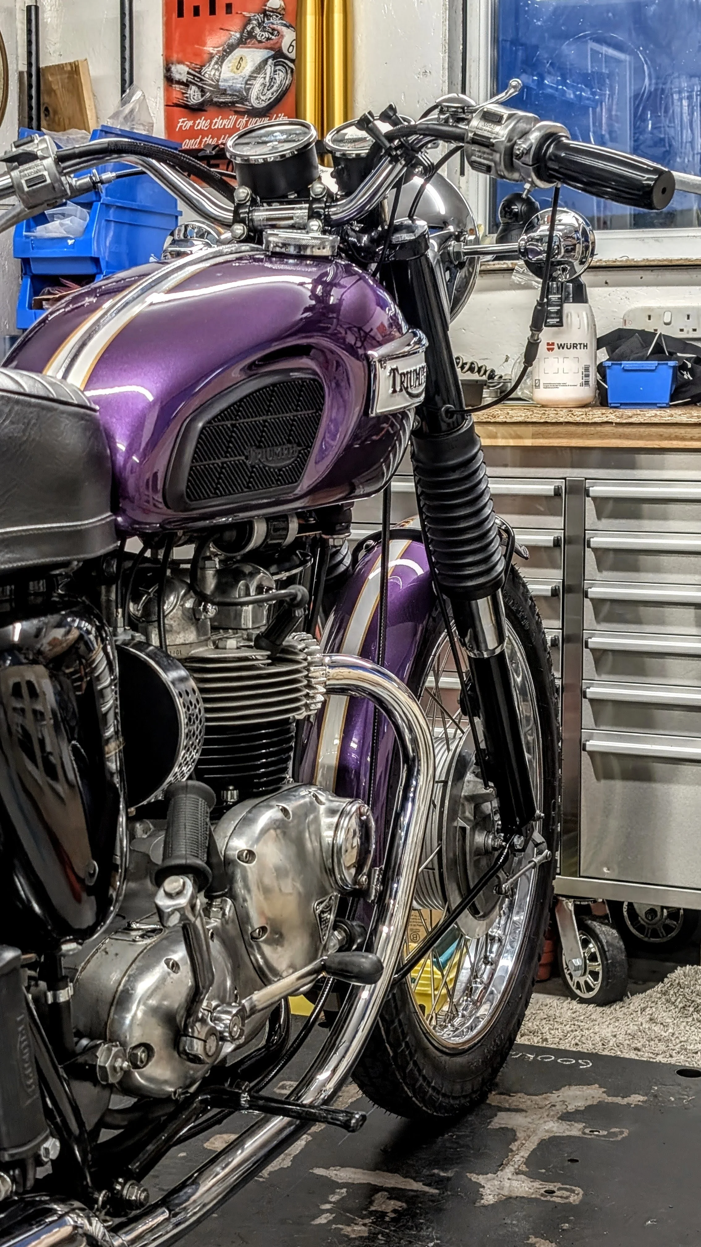 A vintage purple Triumph motorcycle in a workshop with tools and storage cabinets in the background. The bike is just completing a full nut and bolt classic British motorcycle restoration from a barn find condition. 
