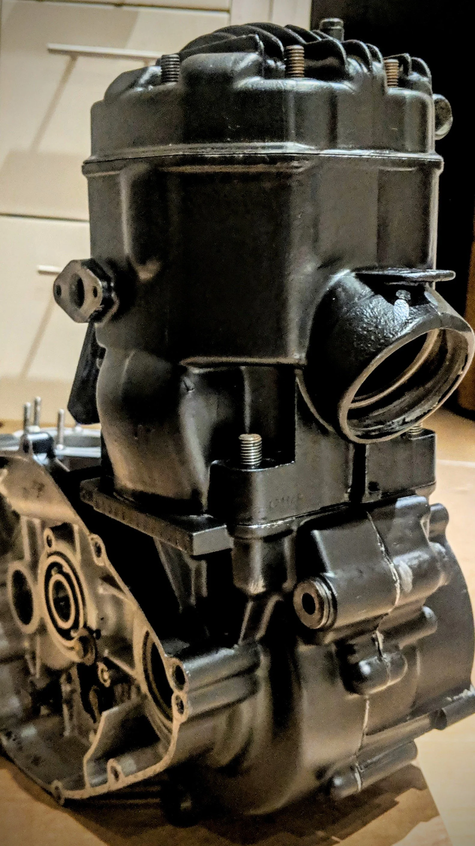 Close-up of a black engine block with visible cylinders and metallic parts, placed on a wooden surface indoors.