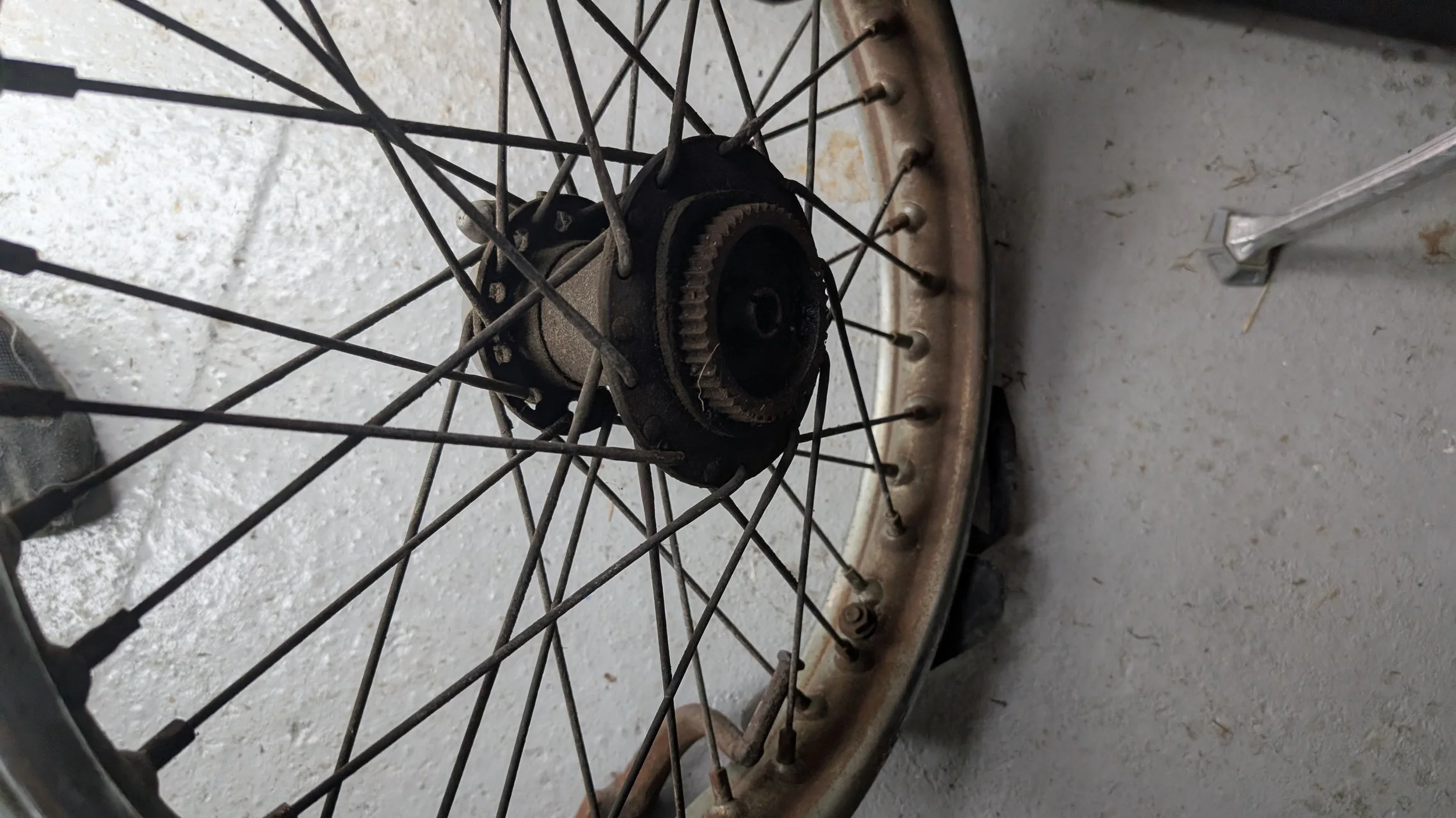 A worn and rusted Triumph T100 QD Quick Disassembly rear wheel with metal spokes and an old hub, resting on a concrete surface. Pitted chrome. Barn find Triumph T100 restoration project. 