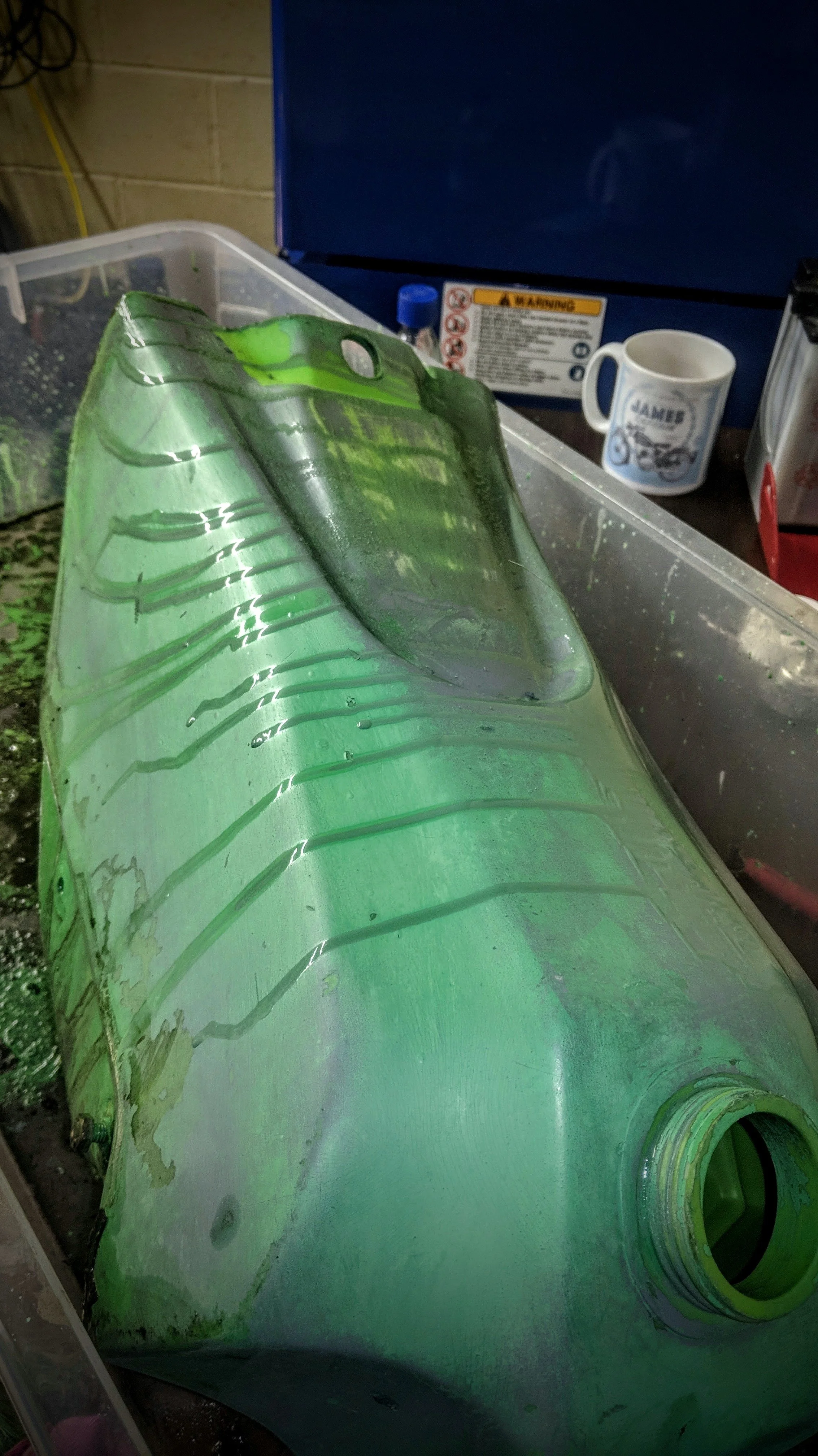 A green plastic bottle lying inside a clear plastic container with a lid. In the background, there is a mug, a blue cabinet, warning labels, and some containers on a workbench.