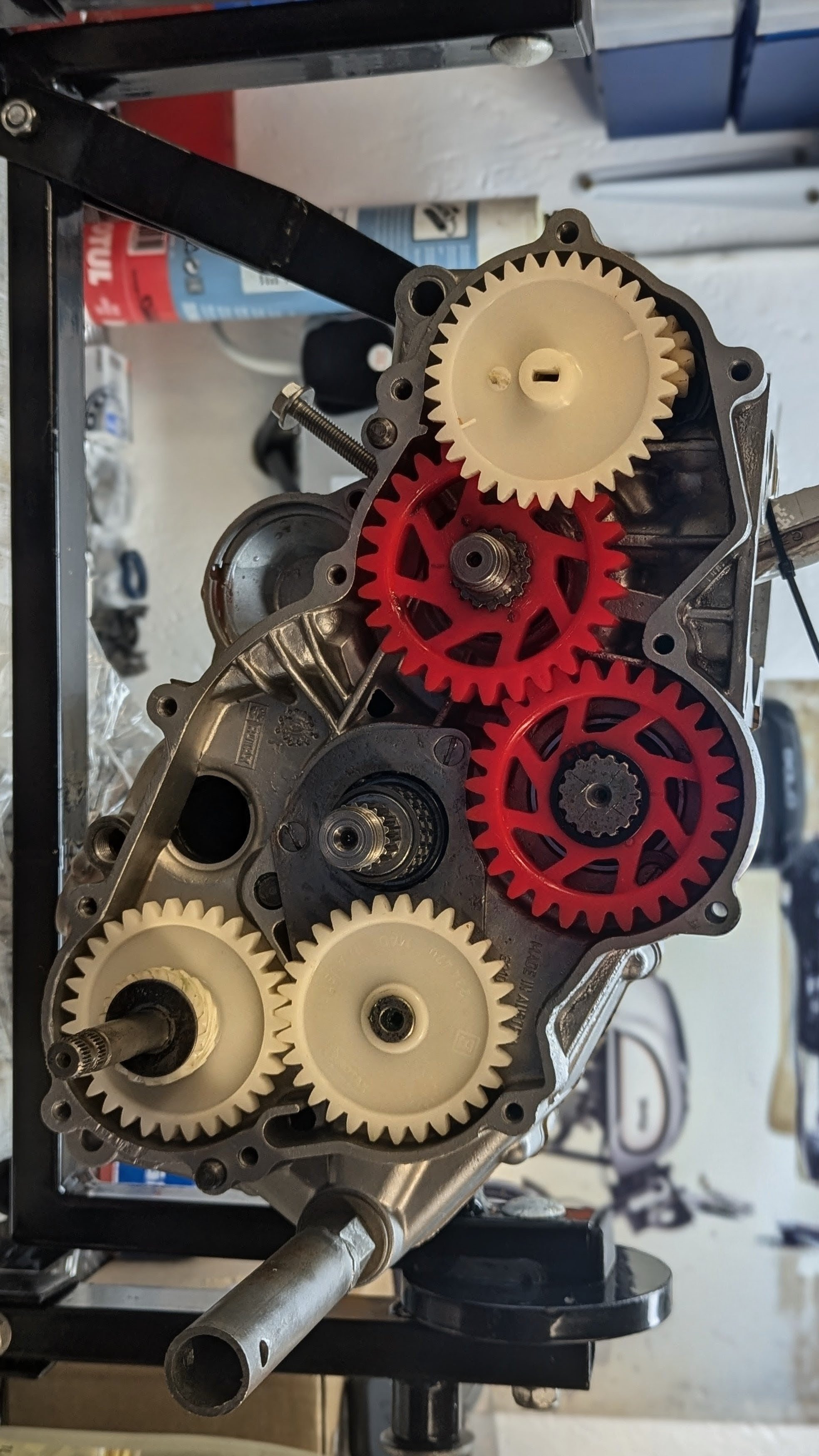 Close-up of a mechanical assembly with white and red gears inside a metal casing, mounted on a black stand or frame.