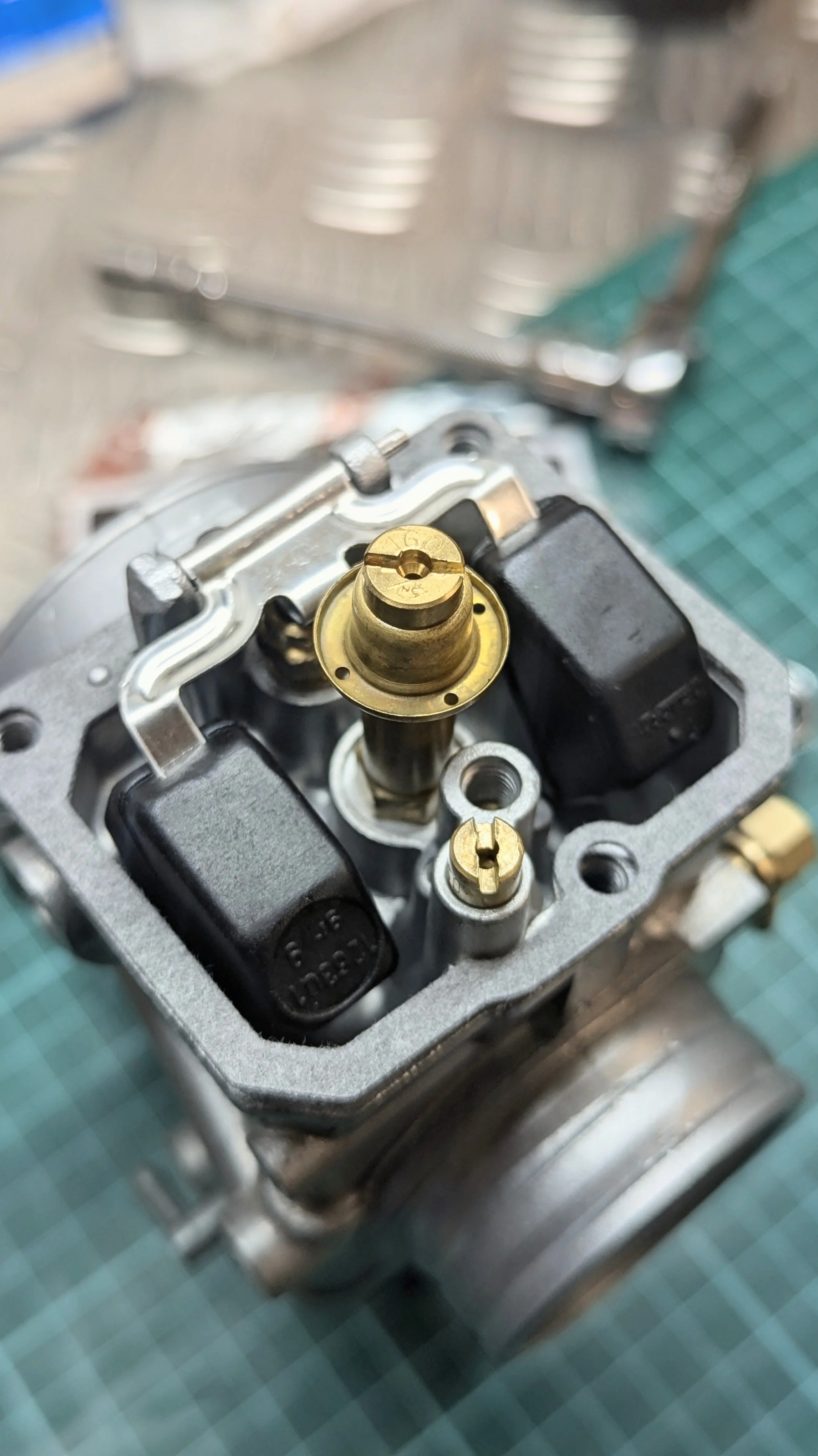 Close-up of a motorcycle cylinder head with brass valve guides, black valve covers, and metal components, on a green work surface.