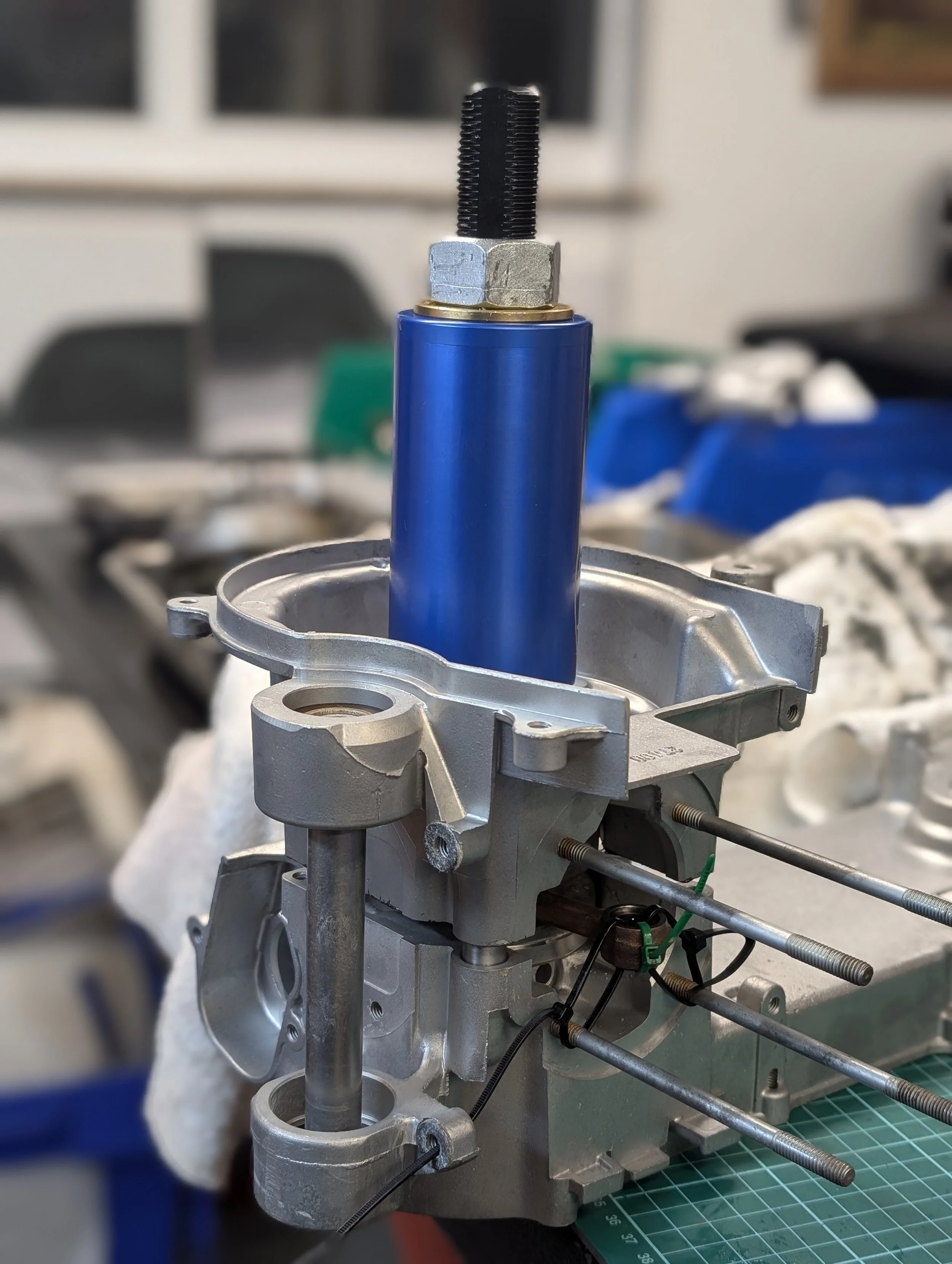 Close-up of mechanical parts including a blue cylinder, metal nuts, bolts, and rods, likely part of machinery or an engine component, with a blurred workshop background.