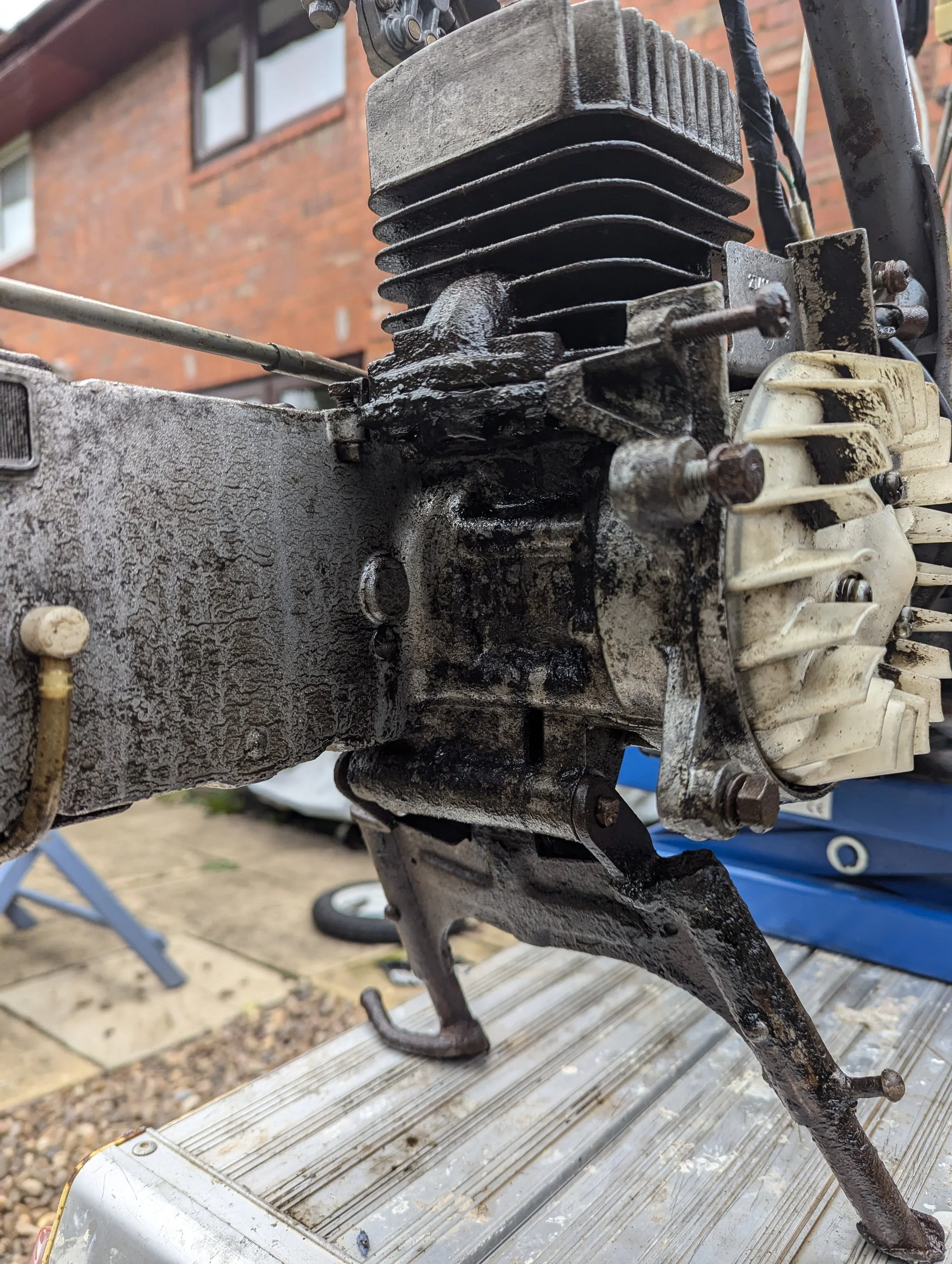Close-up of a rusty, dirty small engine with multiple cooling fins, mounted on a weathered metal surface outdoors with a brick building and other objects in the background.