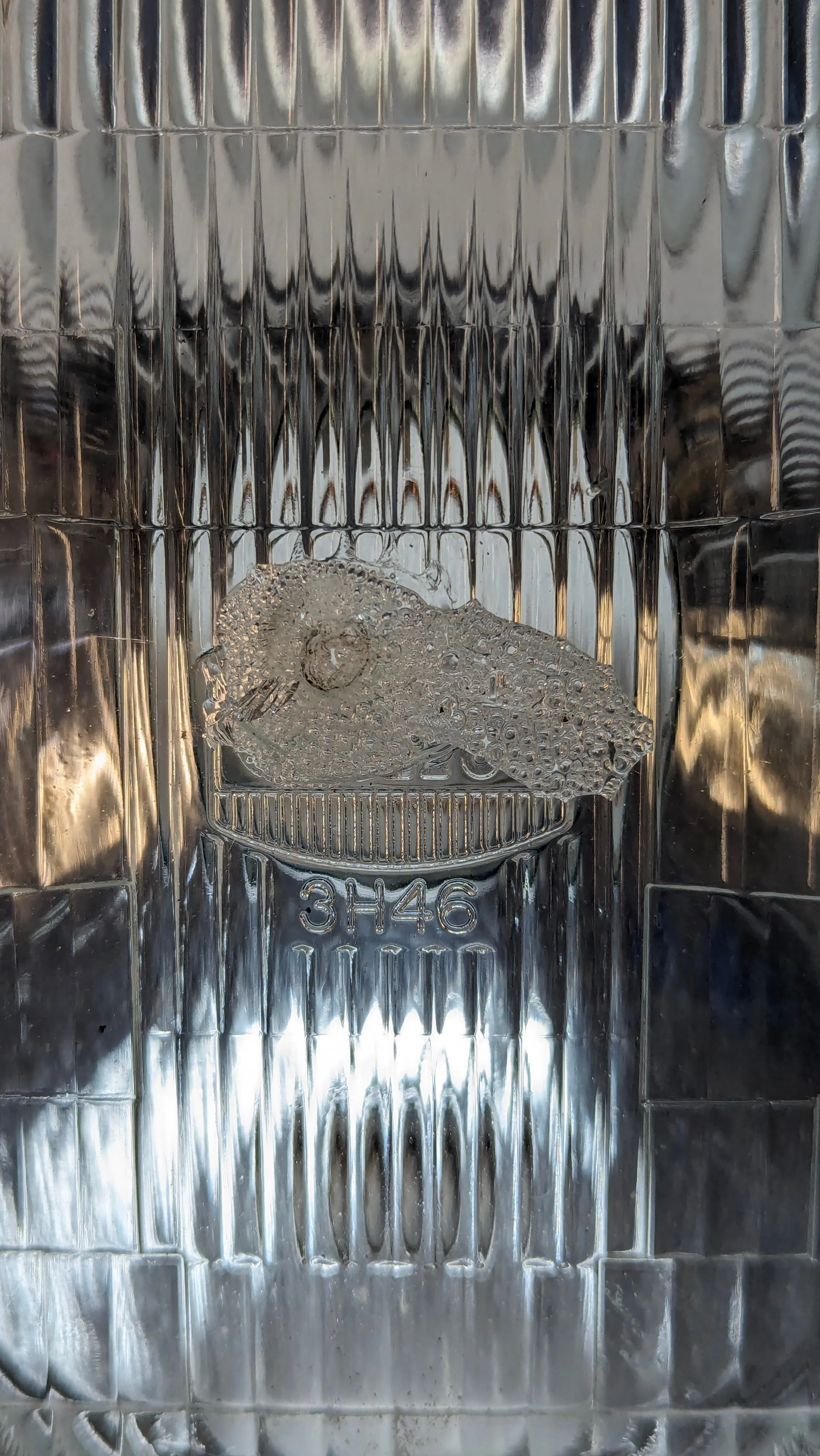 A metal ice cube tray with a partially frozen ice cube inside, marked with the code '3H46' on the bottom.