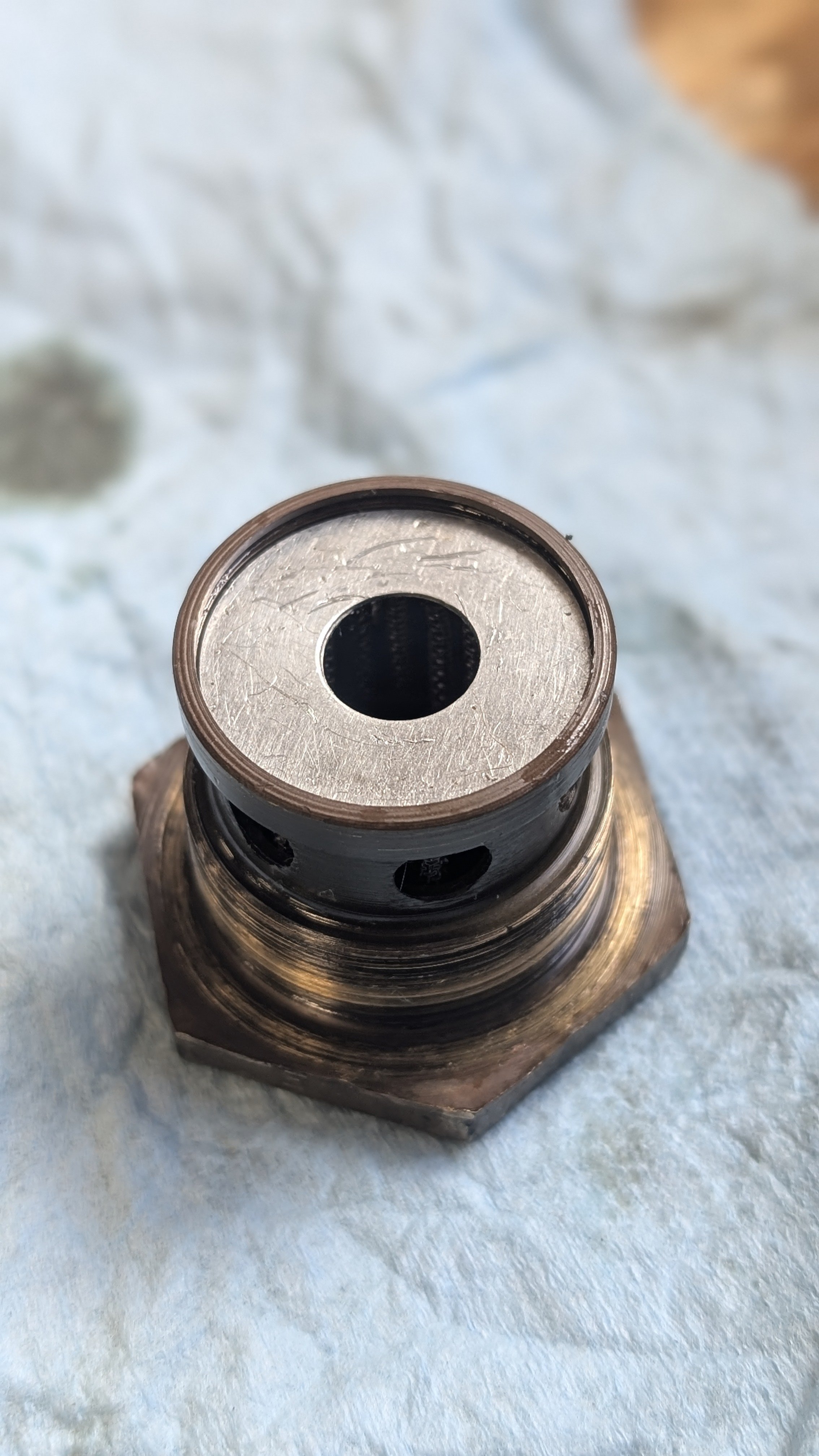 Close-up of a metallic threaded mechanical component, possibly a sensor or a valve, on a light-colored textured surface.