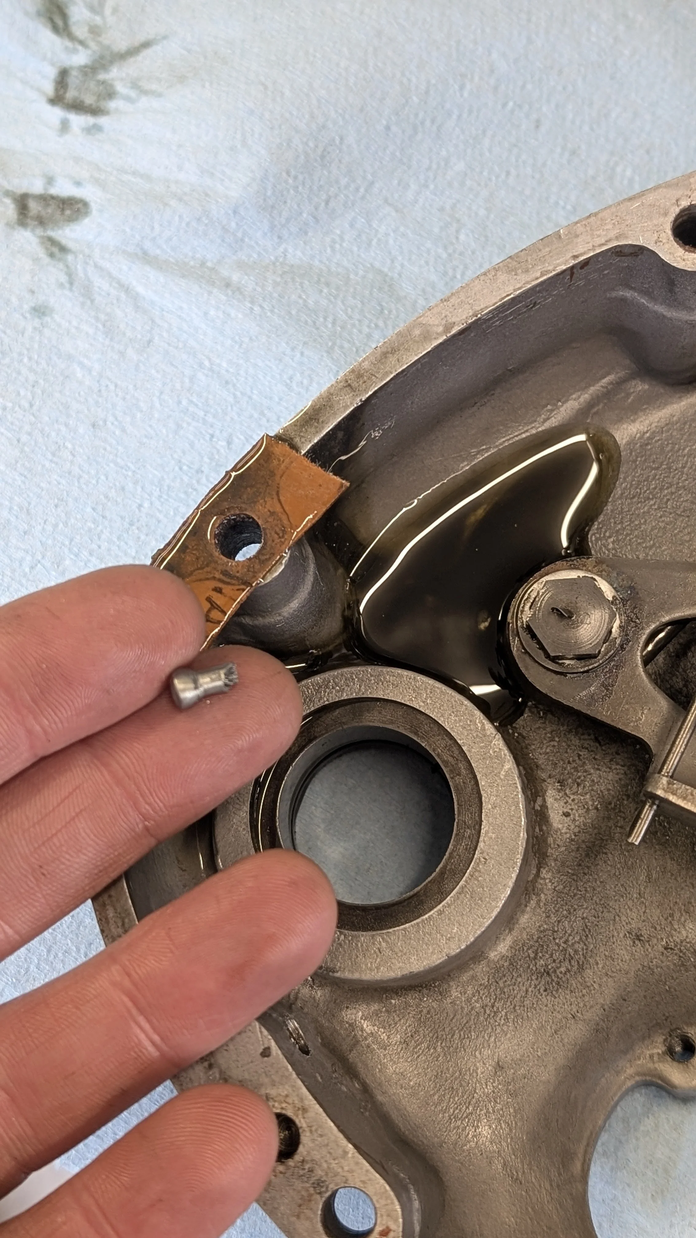 Close-up of a person inspecting or working on a metal engine part, possibly a cylinder head or similar component, with tools and a small metal piece in hand.