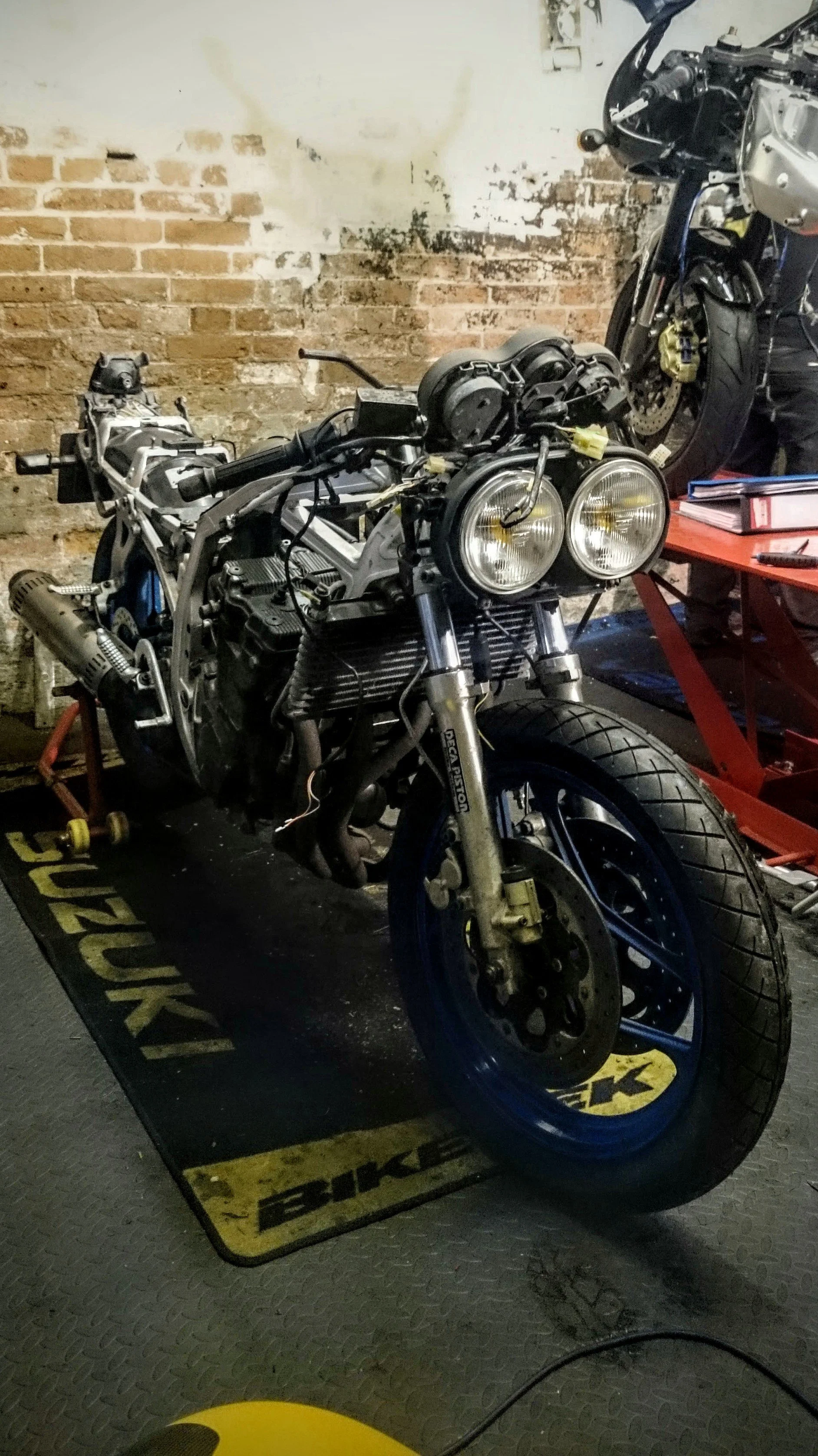 A motorcycle on a work stand in a garage, partially disassembled with visible engine parts, in front of a brick wall.