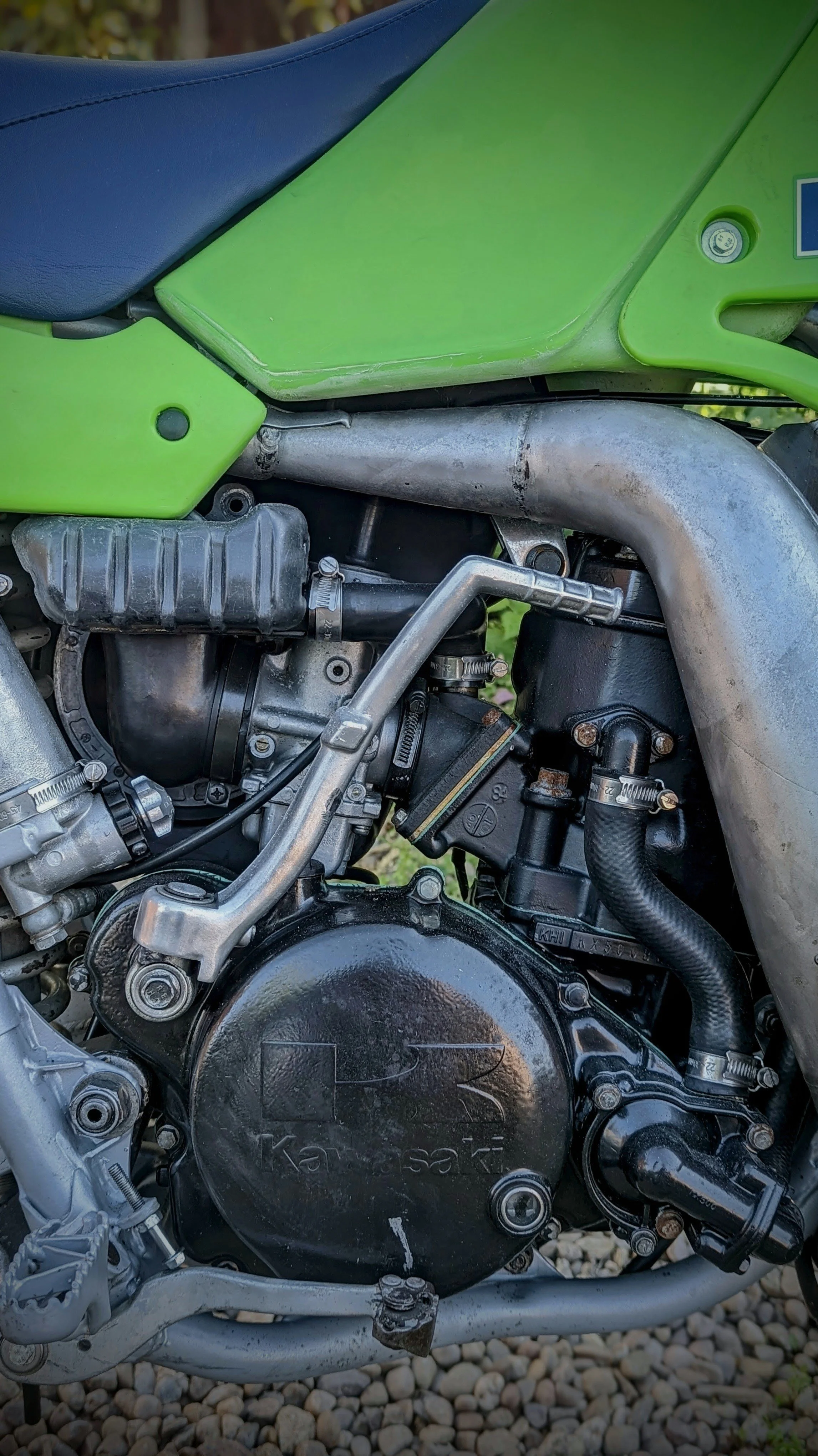 Close-up of a green Kawasaki motorcycle engine with various metal parts, hoses, and a black cover, resting on a bed of small rocks.