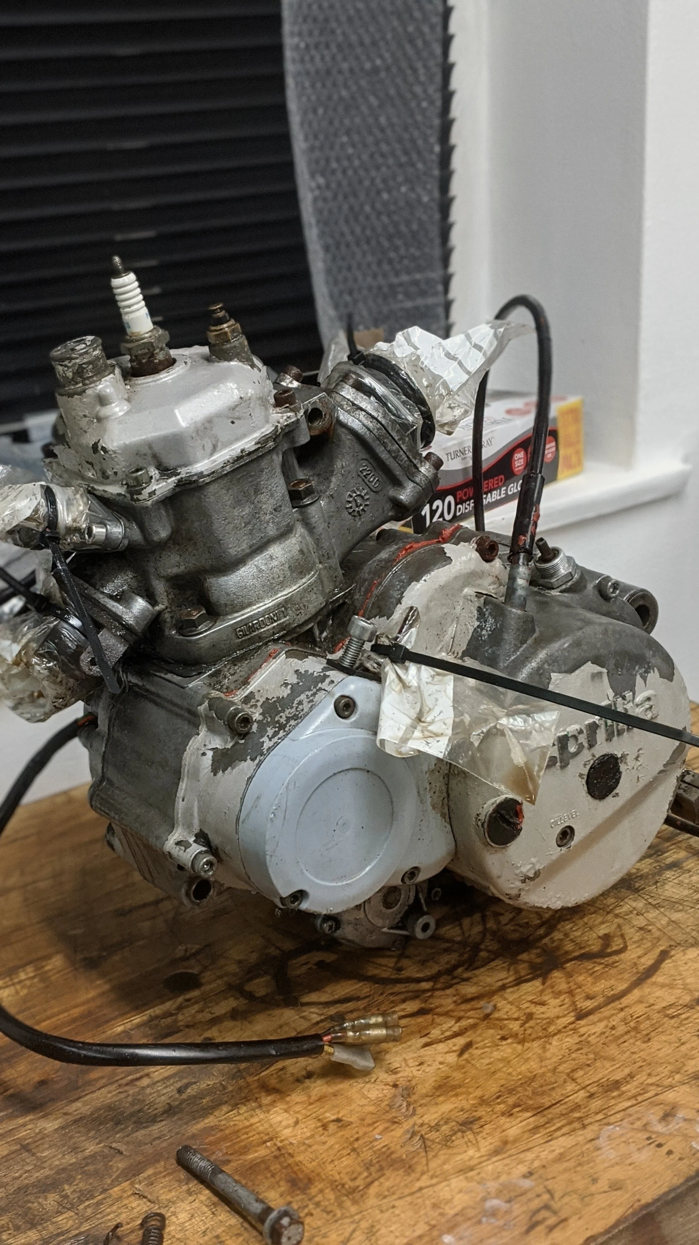 A disassembled motorcycle engine placed on a wooden surface with tools and wires around it.