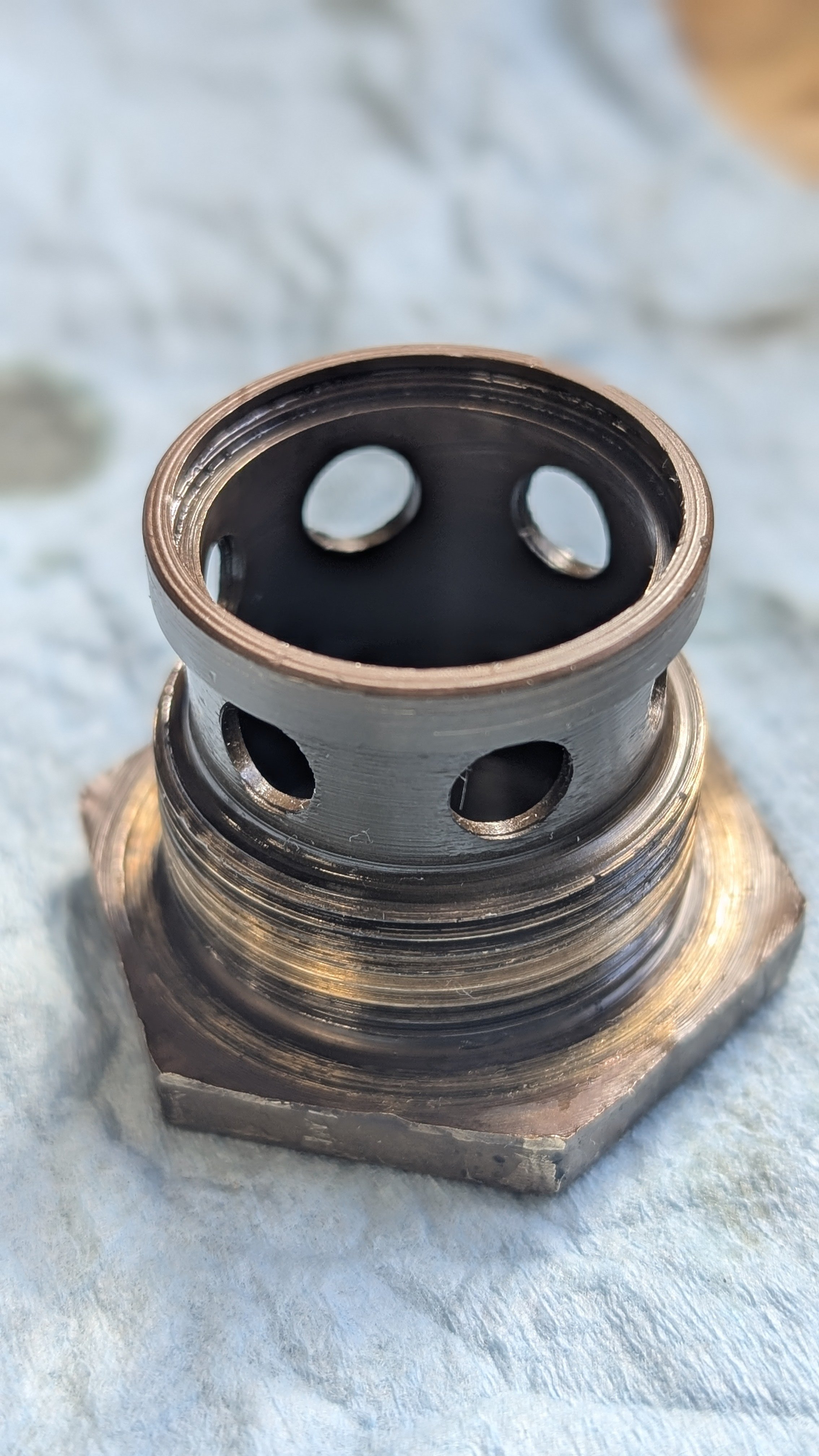Close-up of a metallic mechanical part with multiple holes and a threaded section, placed on a light-colored textured surface.