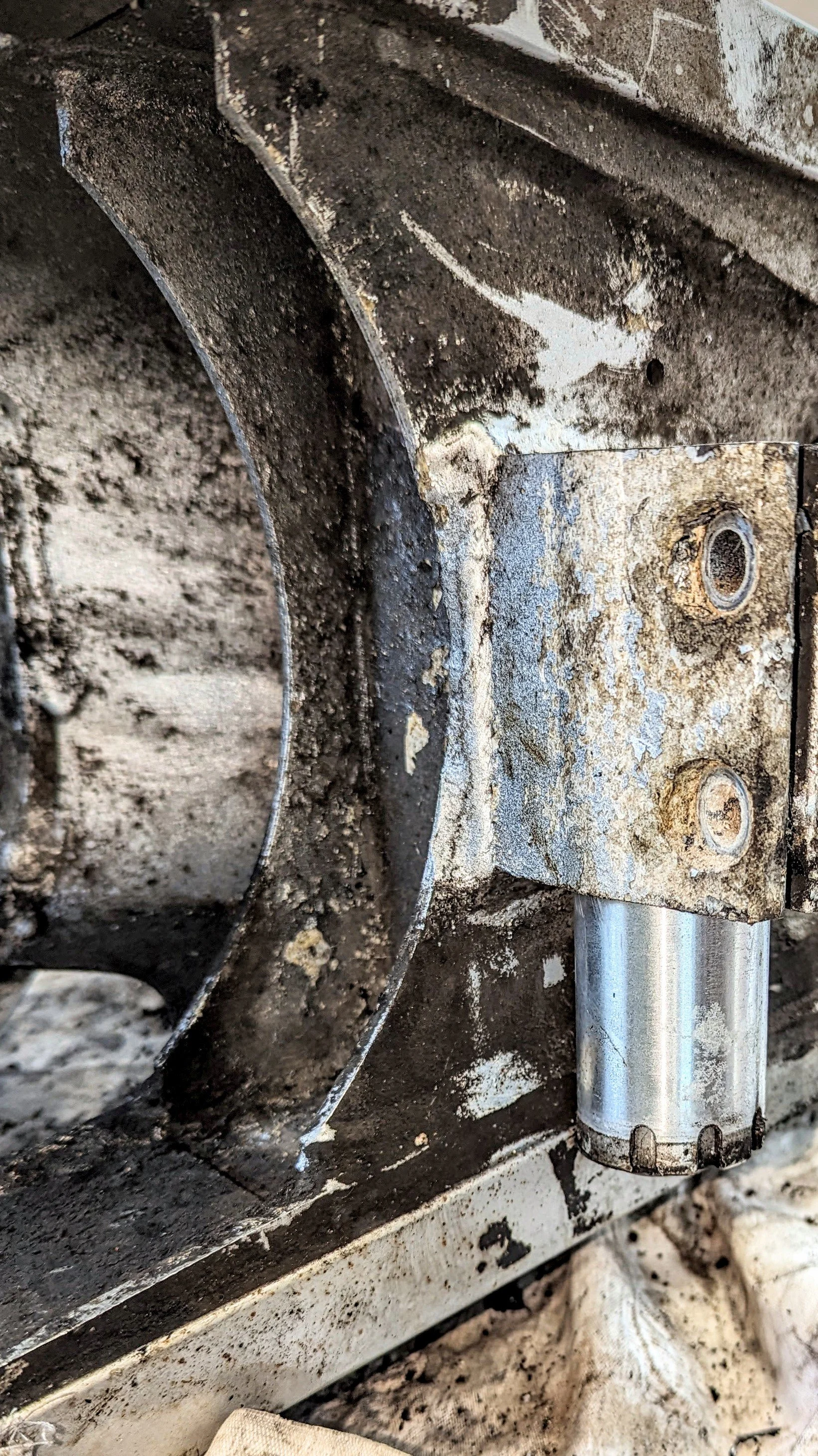 Close-up of a rusty, dirty mechanical part on a machine or vehicle, with visible screws, a metal rod, and dirt surrounding it.