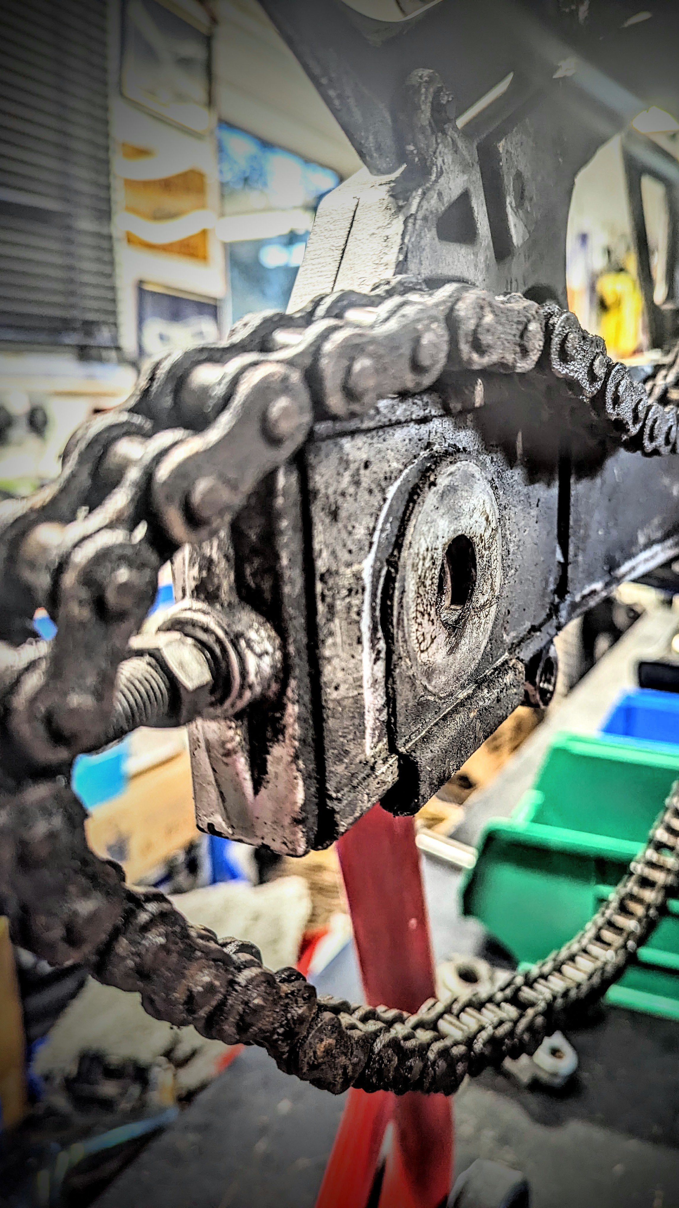 Close-up of a rusty, dirty chain and sprocket on a mechanical device or machine, with a blurry workshop or garage background.
