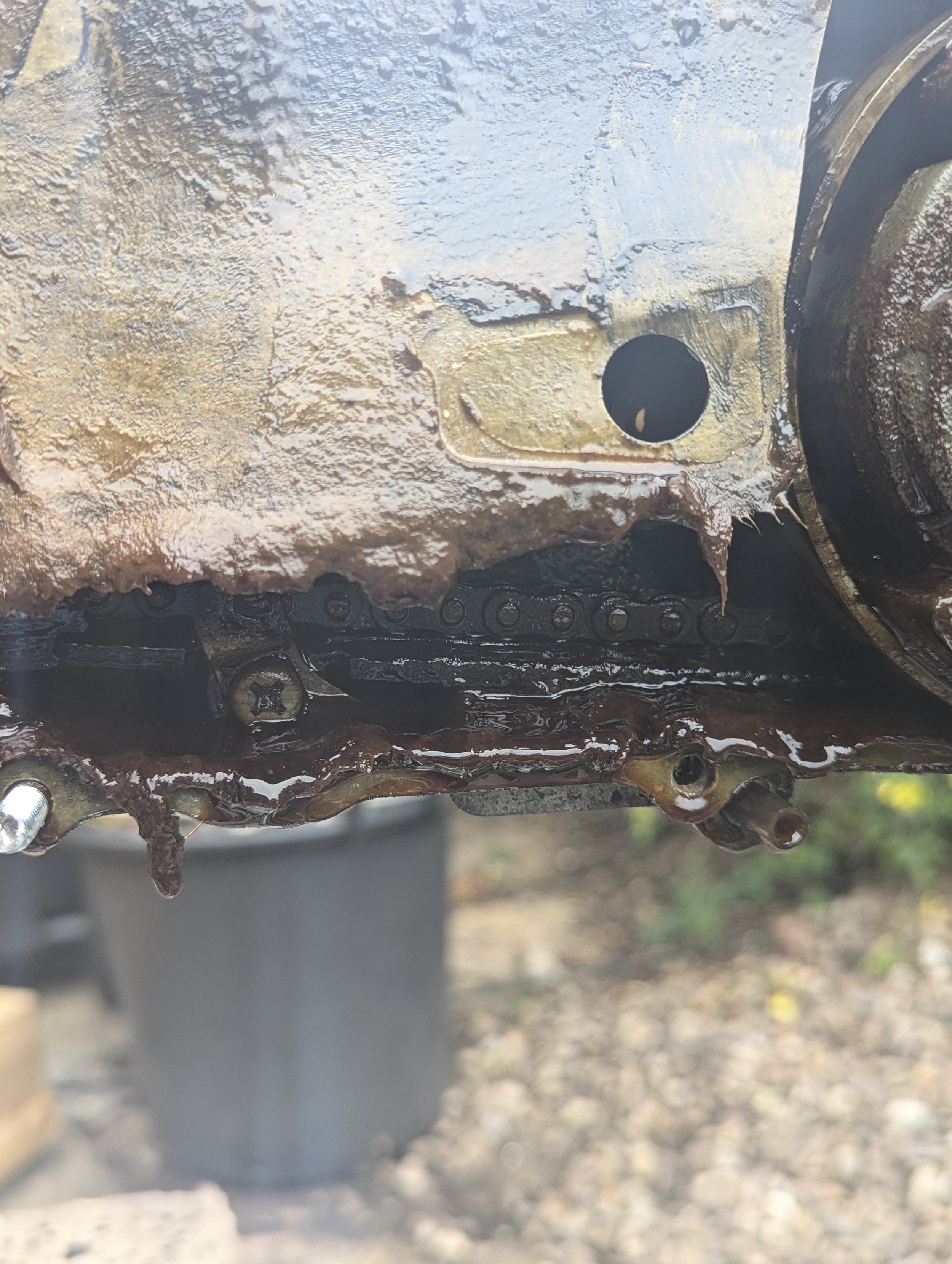 Close-up photo of a rusty, oil- and grime-covered mechanical part, possibly an engine component, with a background of grass and dirt.