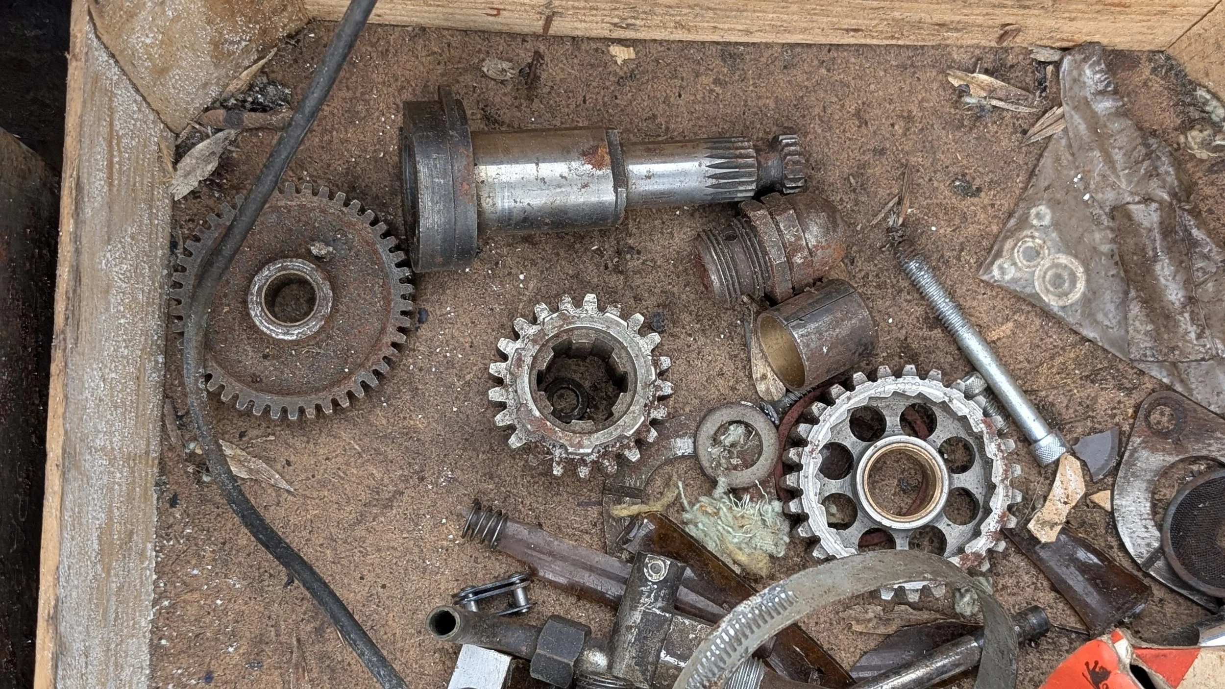 Disorganized mechanical parts including gears, shafts, screws, and metal debris scattered on a dirty work surface. Barn find Triumph T100 gearbox parts for restoration project. 