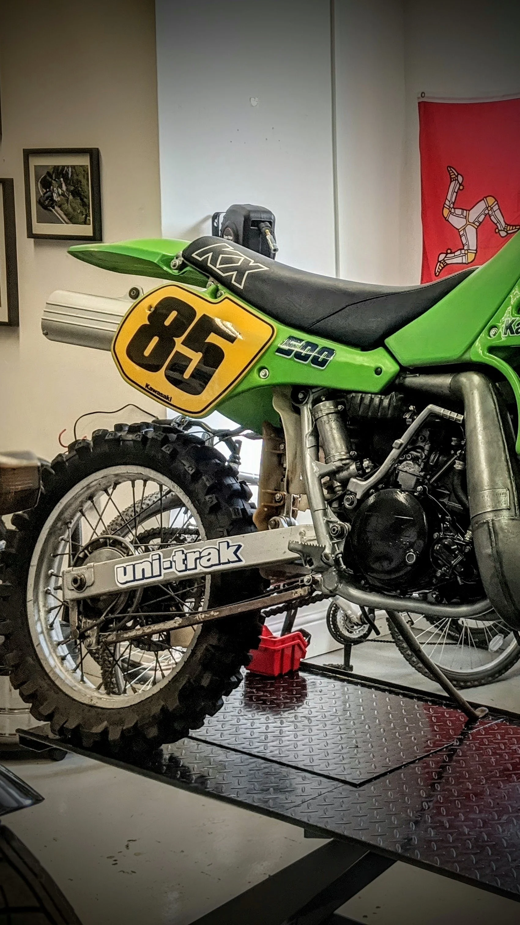 A partially disassembled green Kawasaki dirt bike with a yellow number plate '85', resting on a metal work surface, in a workshop with various framed pictures and a red poster in the background.