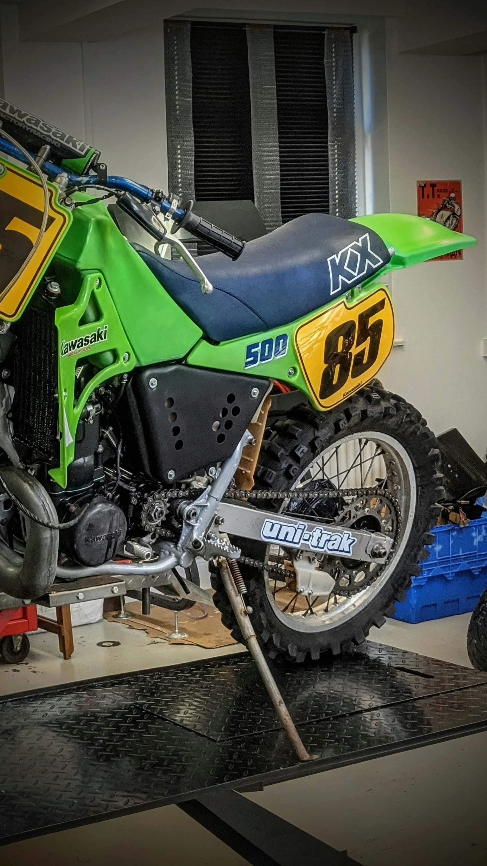 A green Kawasaki motocross bike with number 845 on the side, positioned on a work stand in a garage. The bike's seat is black with white logos, and the background shows a window with blinds and various tools and storage containers around.