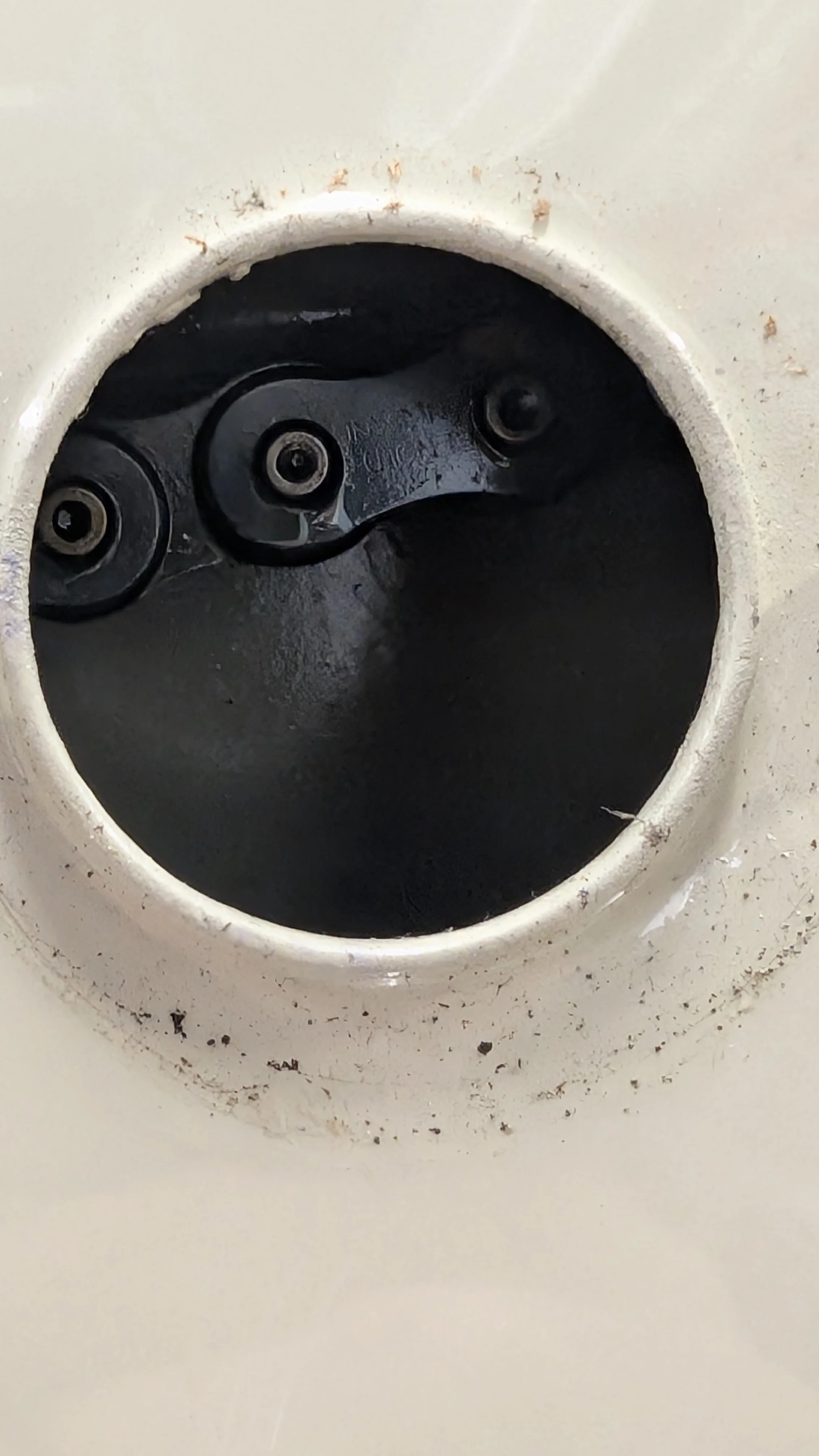 Close-up view of a circular opening in a white surface, revealing internal black components with screws, and some dirt around the edges.