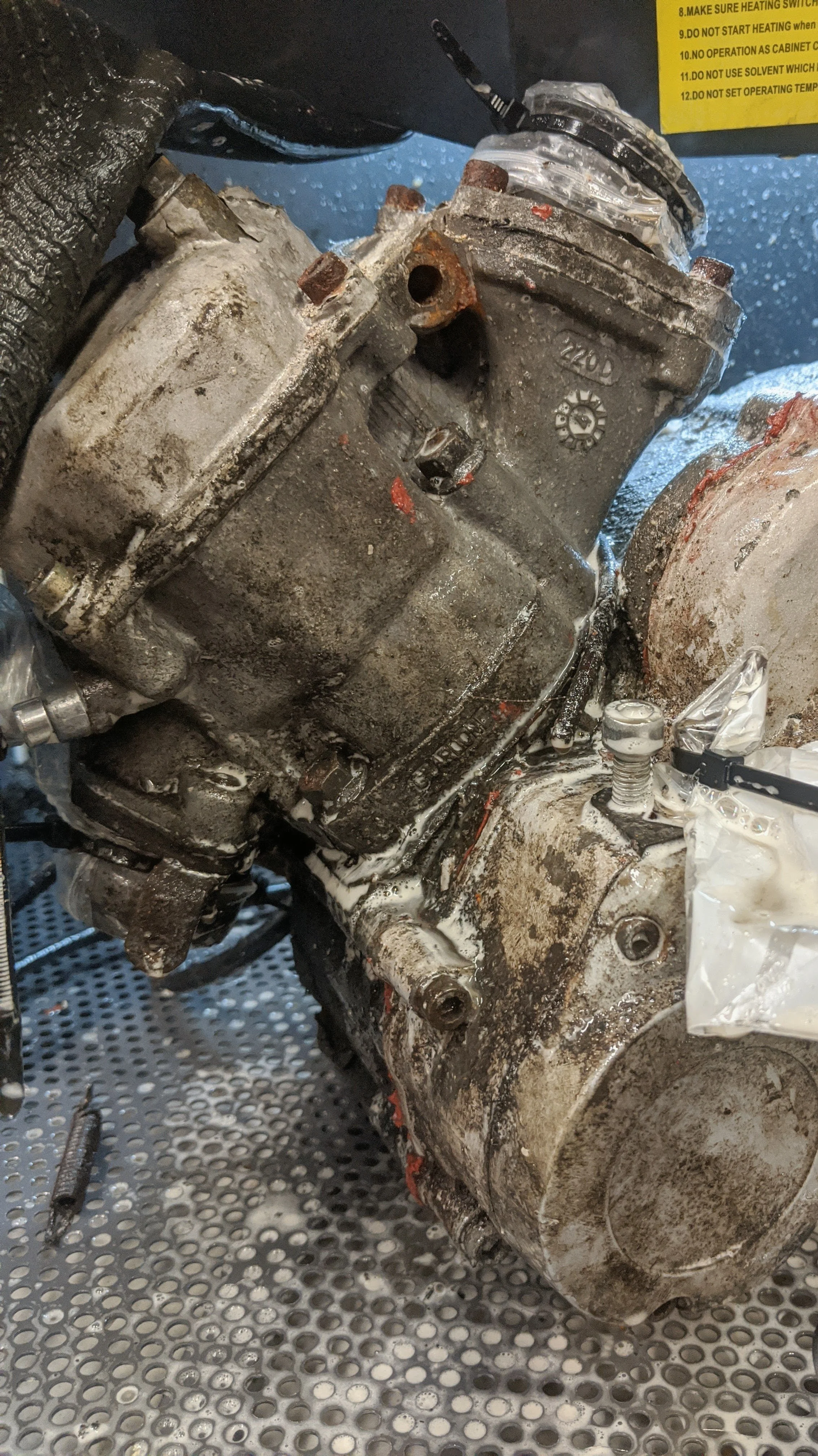 Close-up photo of a dirty and oily engine, with various bolts and components, on a perforated metal surface, with scattered tools and parts around.