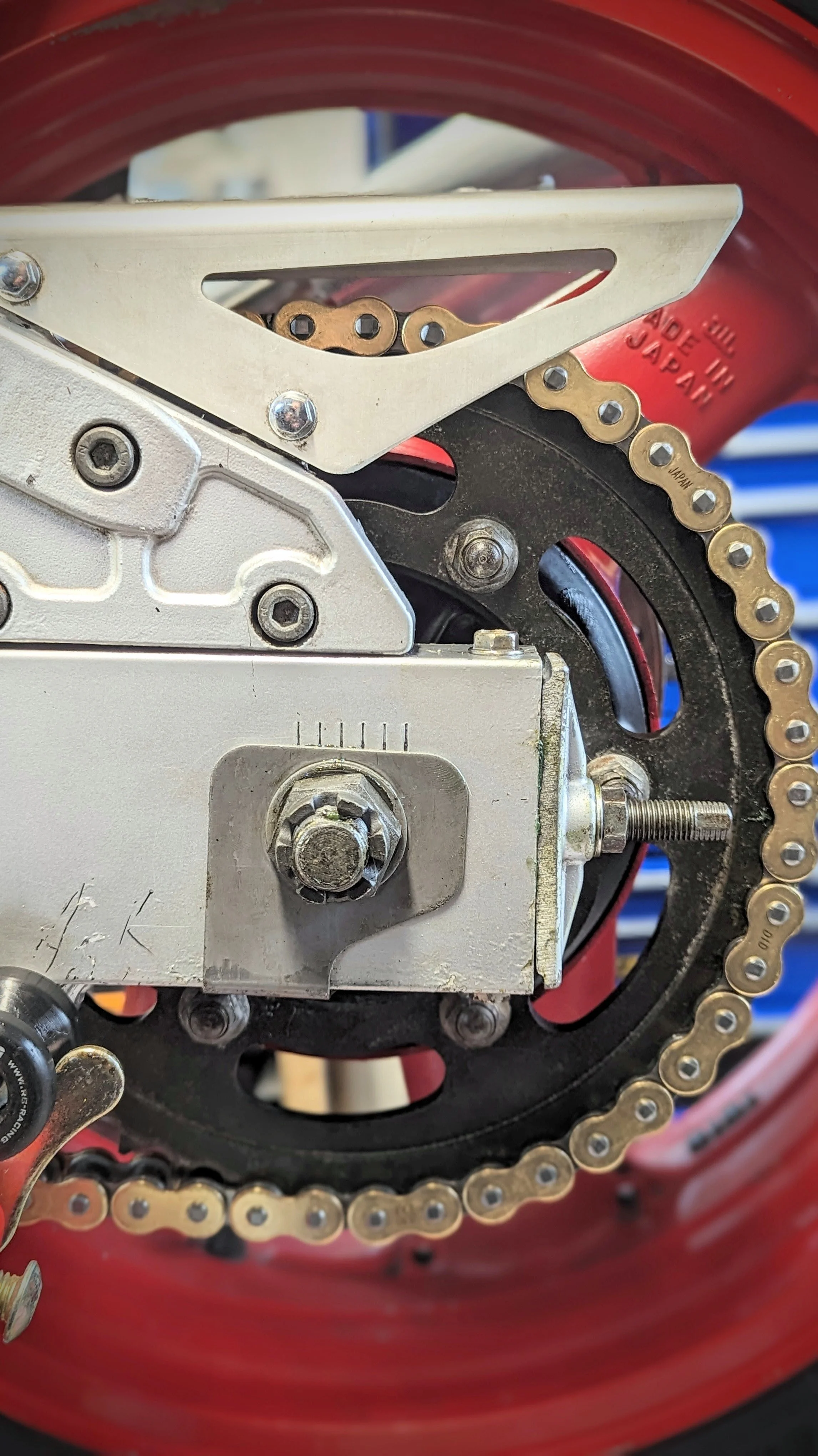 Close-up of a motorcycle chain and sprocket attached to a part of the motorcycle's transmission system, with a red wheel rim in the background.