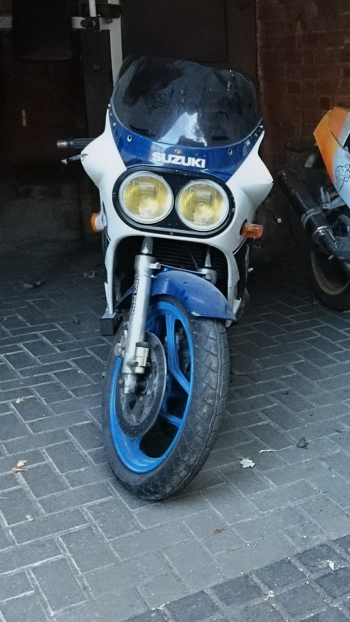 A vintage blue and white Suzuki motorcycle with a black windshield and yellow headlights parked on a brick sidewalk.