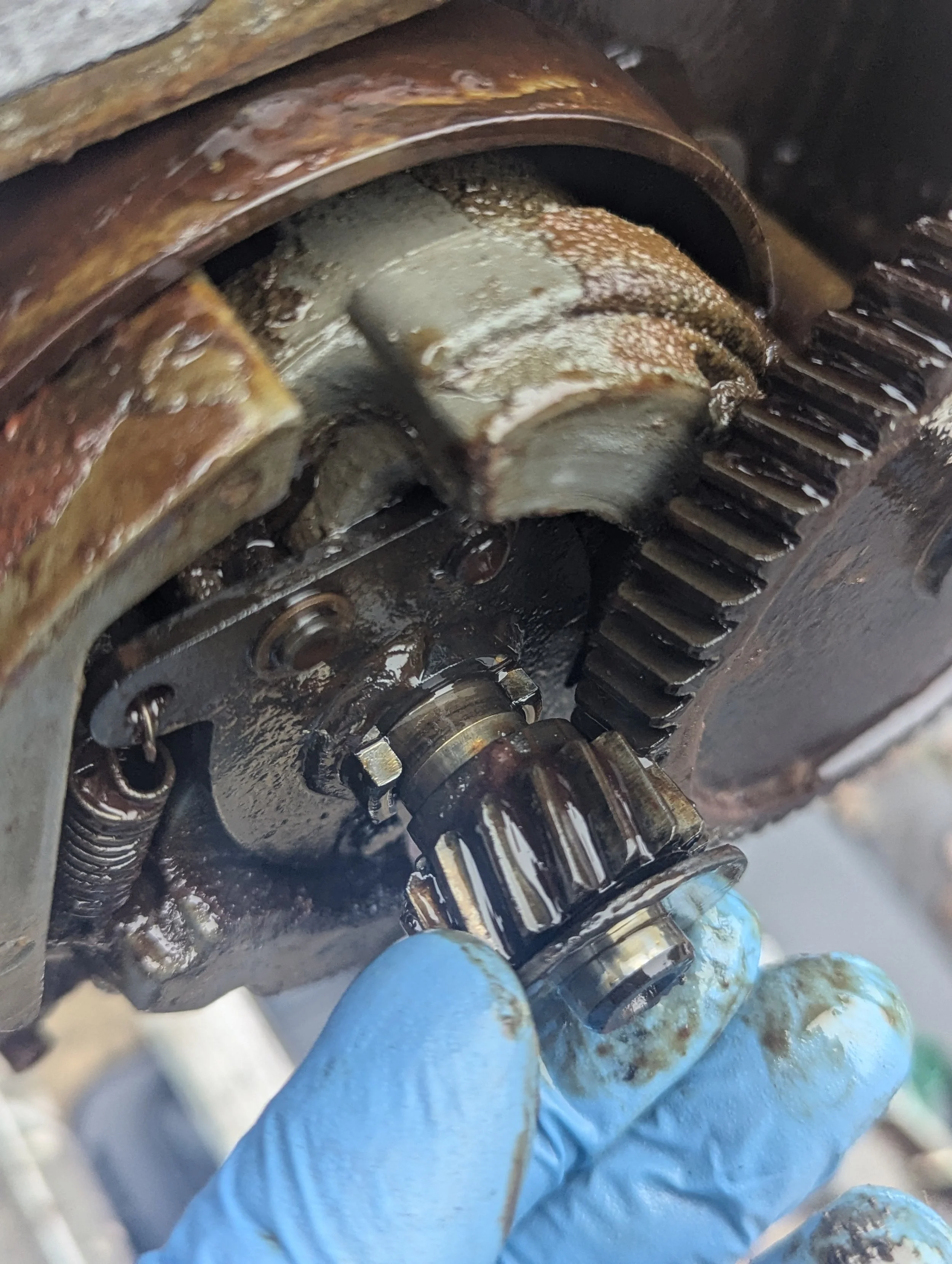 Close-up of a mechanic's gloved hand working on a rusty metal gear component, possibly in an engine or machinery repair setting.