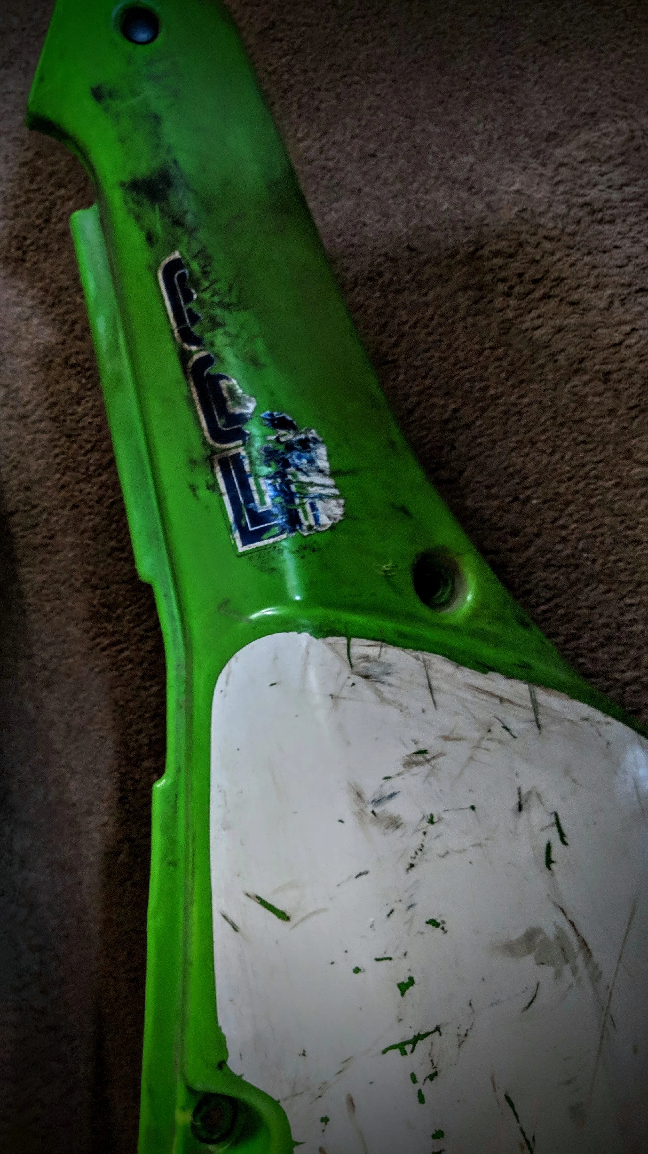 A green dirt bike fender with scratches, dirt, and a partially torn sticker on it, resting on a carpeted floor.