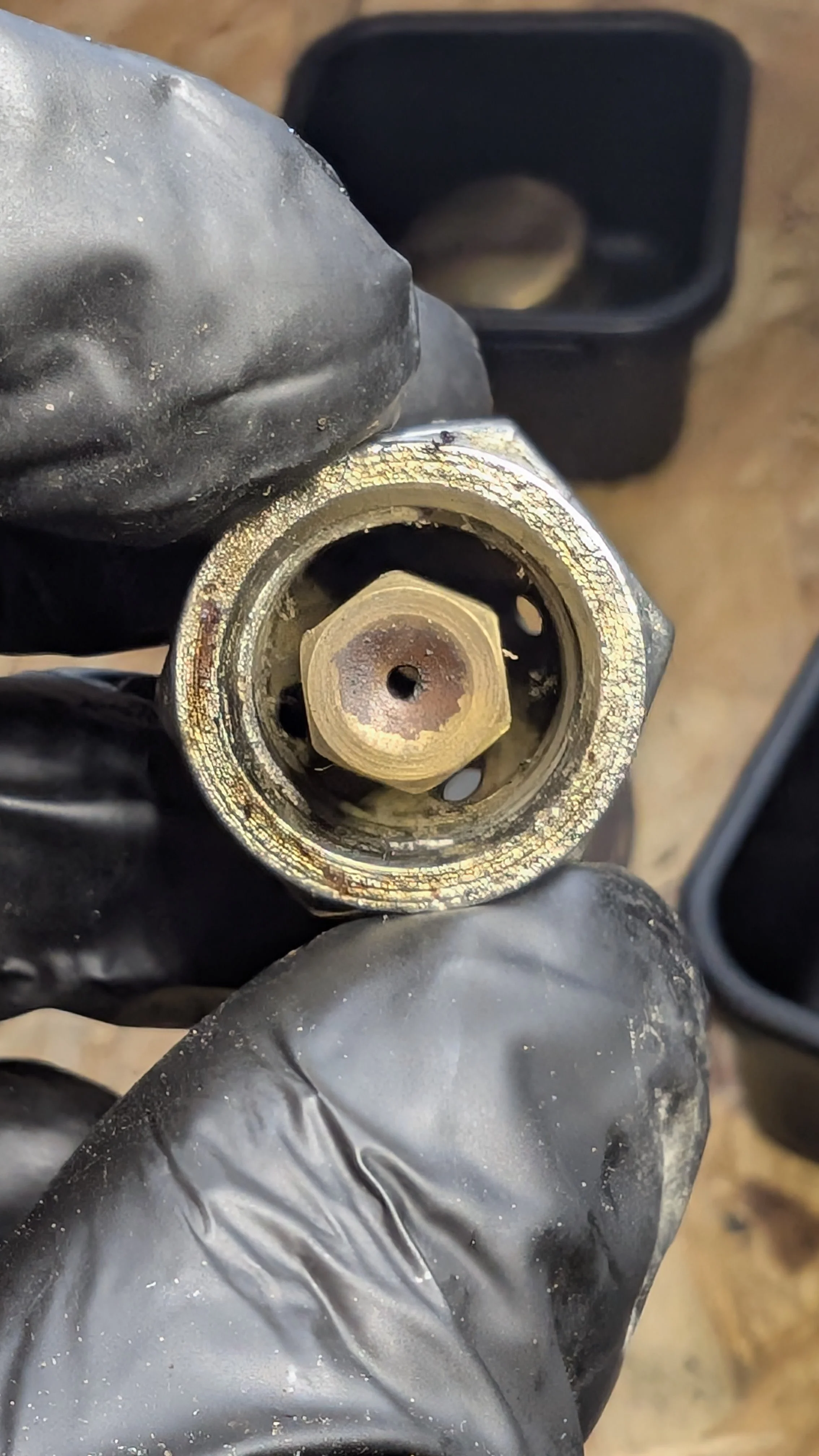 Close-up of a metallic fitting or connector, being held by a person wearing black gloves. There is a black container or device in the background.