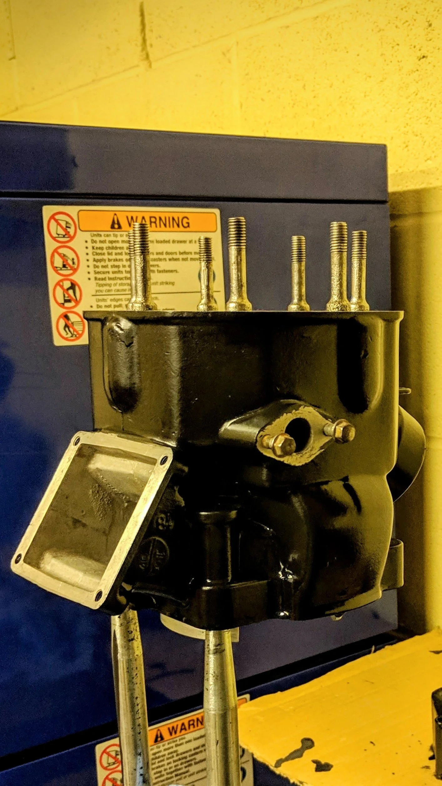 Close-up of an industrial machine with metal pipes protruding from a black metal housing, warning labels, and a yellow work surface with tools.