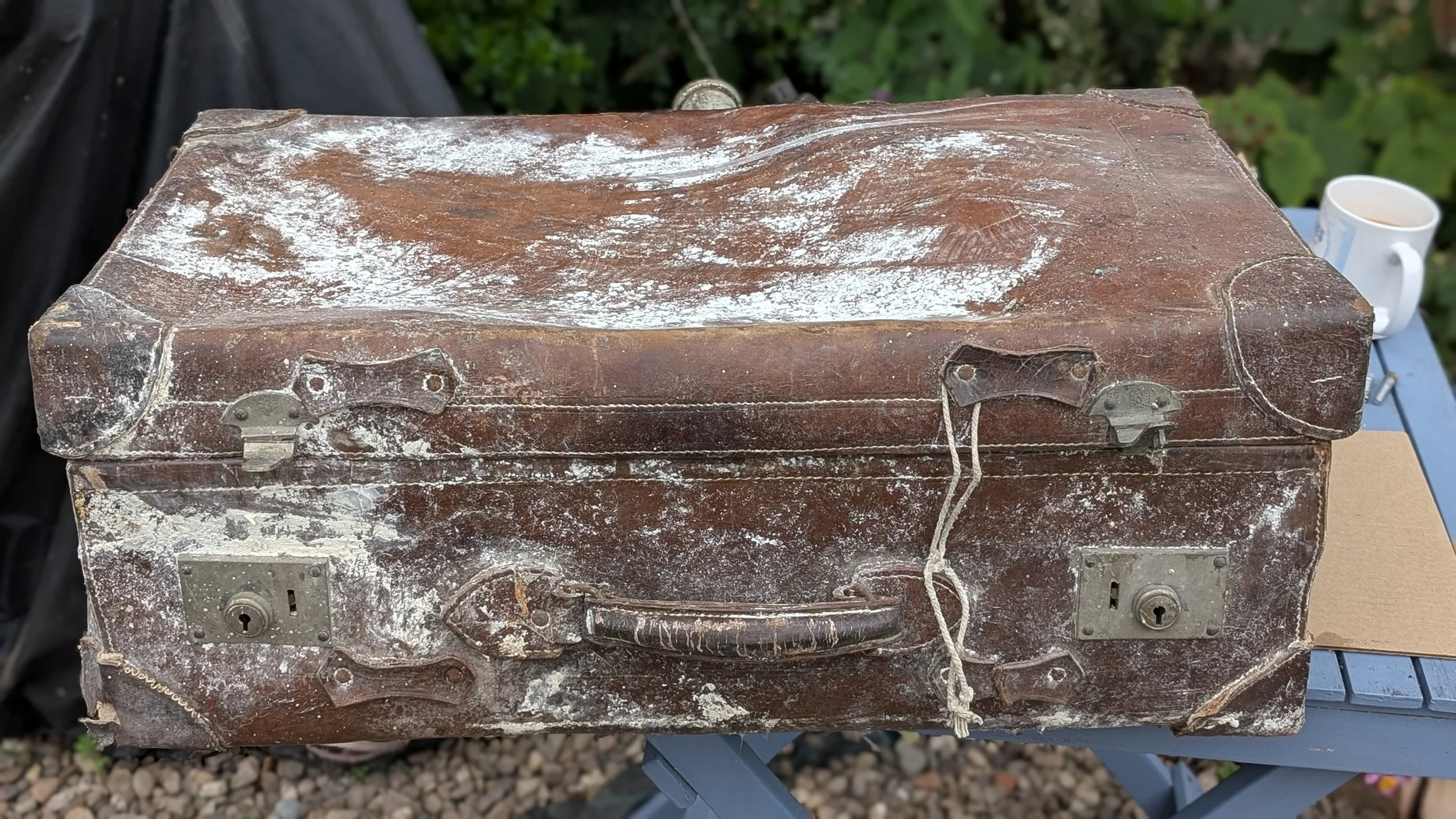 Old, weathered leather suitcase with metal latches, sitting on a blue table outdoors, with a white mug and green foliage in the background.