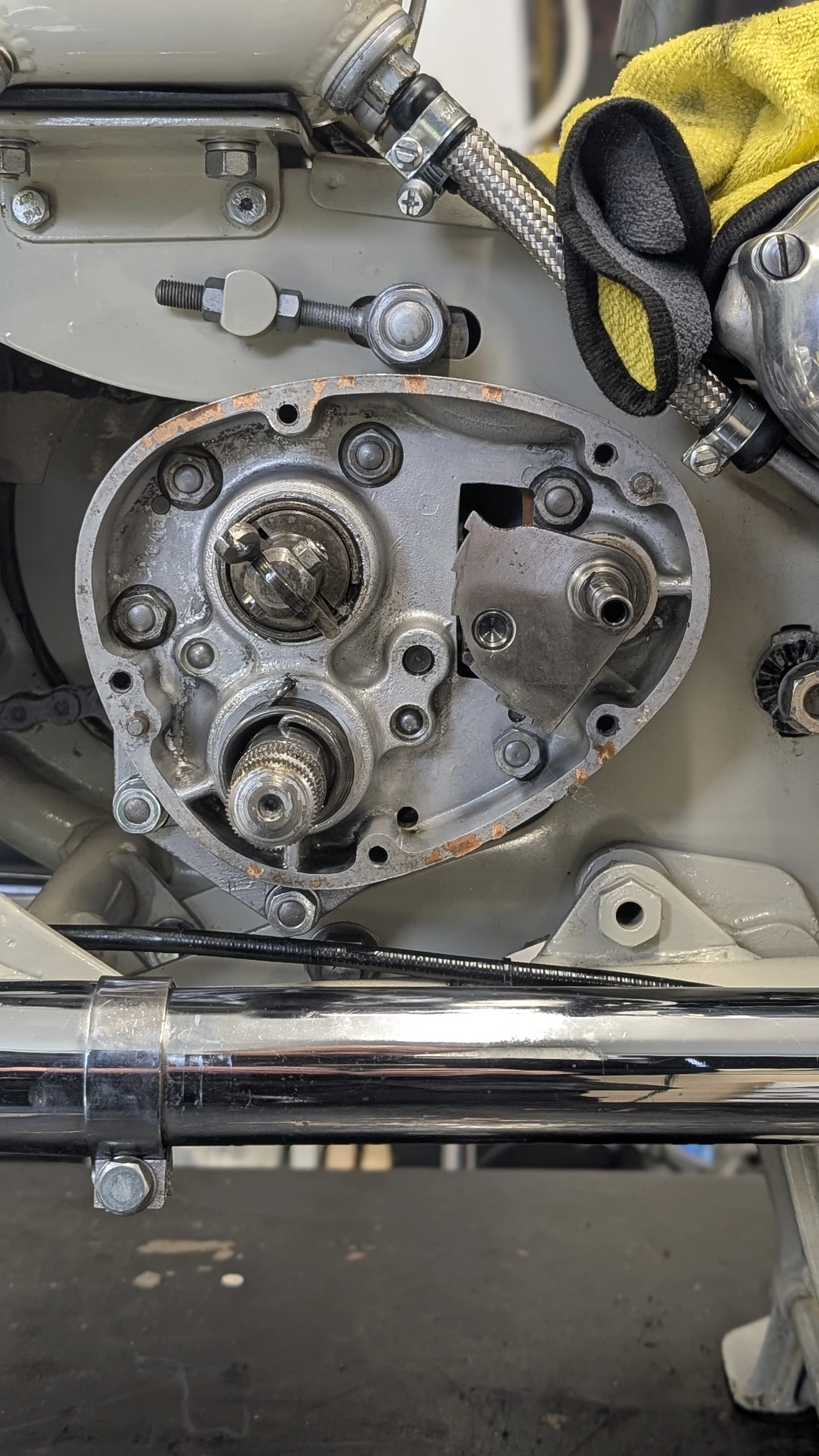Close-up view of a motorcycle engine with the top cover removed, showing internal gears, shafts, and components, with a yellow cloth and braided hose nearby.