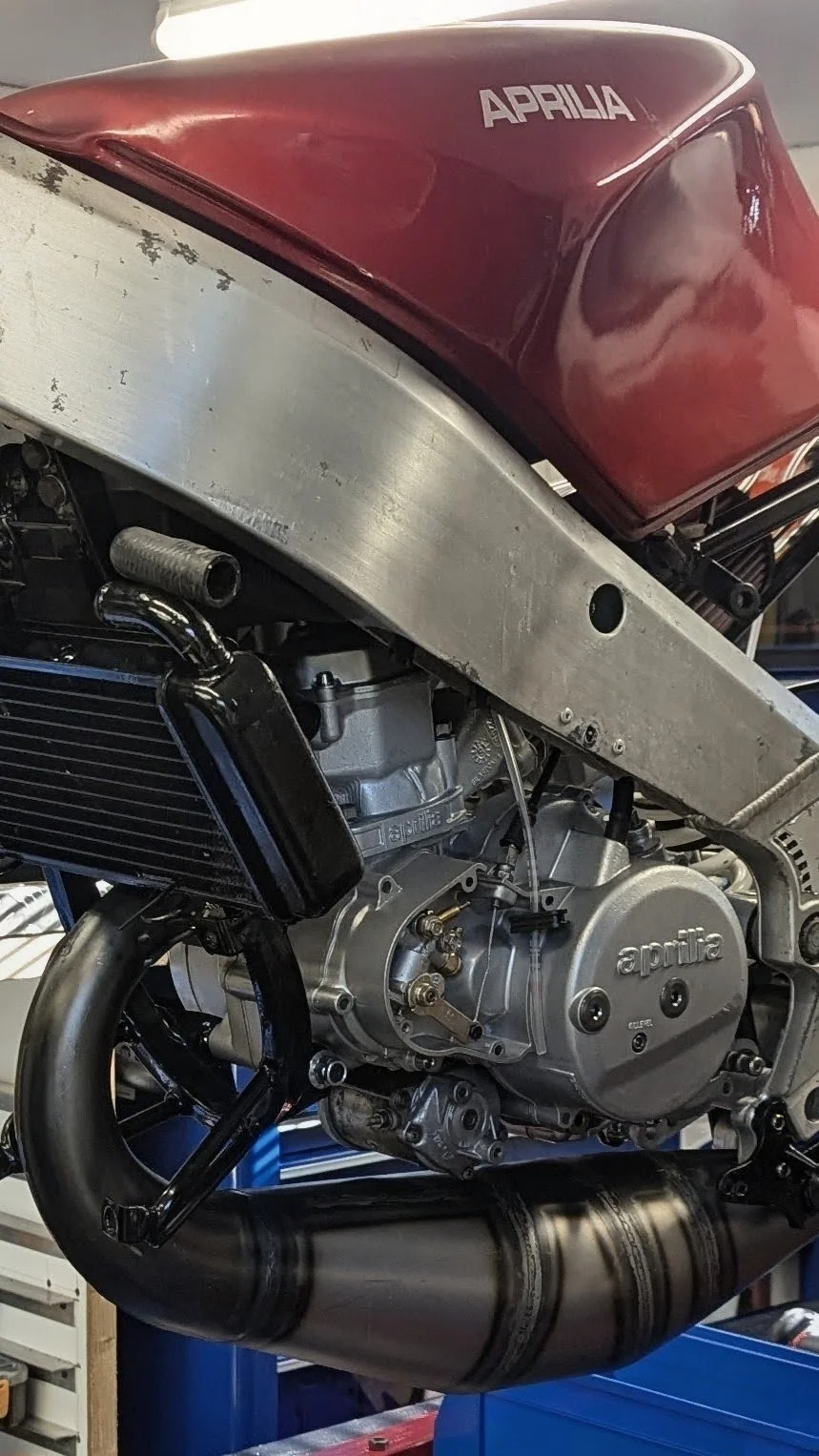 Close-up of an Aprilia motorcycle engine and red fuel tank labeled 'Aprilía' in a workshop.