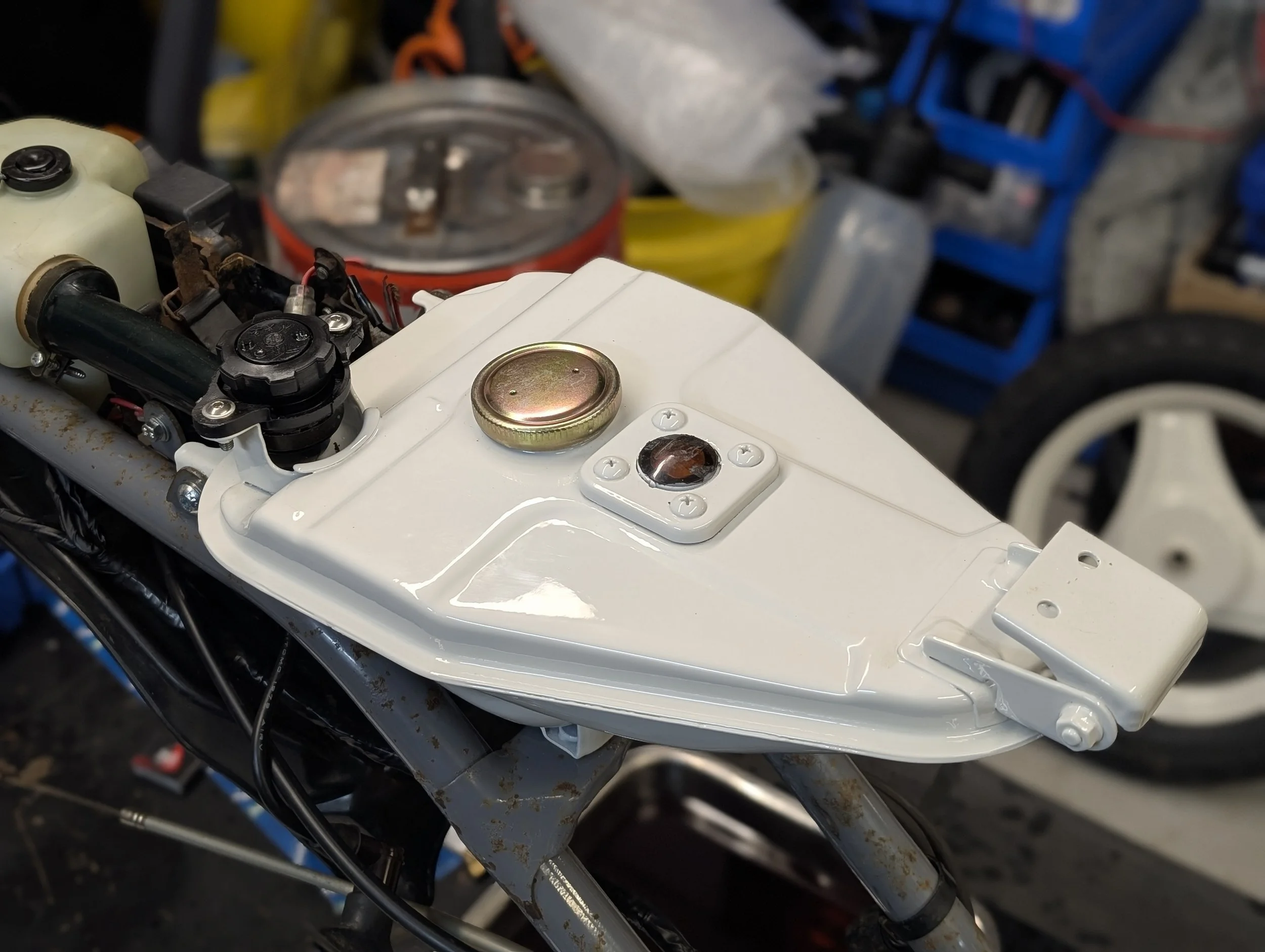 Close-up view of a motorcycle's front section, showing part of the white frame, a gold-colored cap, and nearby wiring, with tools and storage containers in the blurred background.