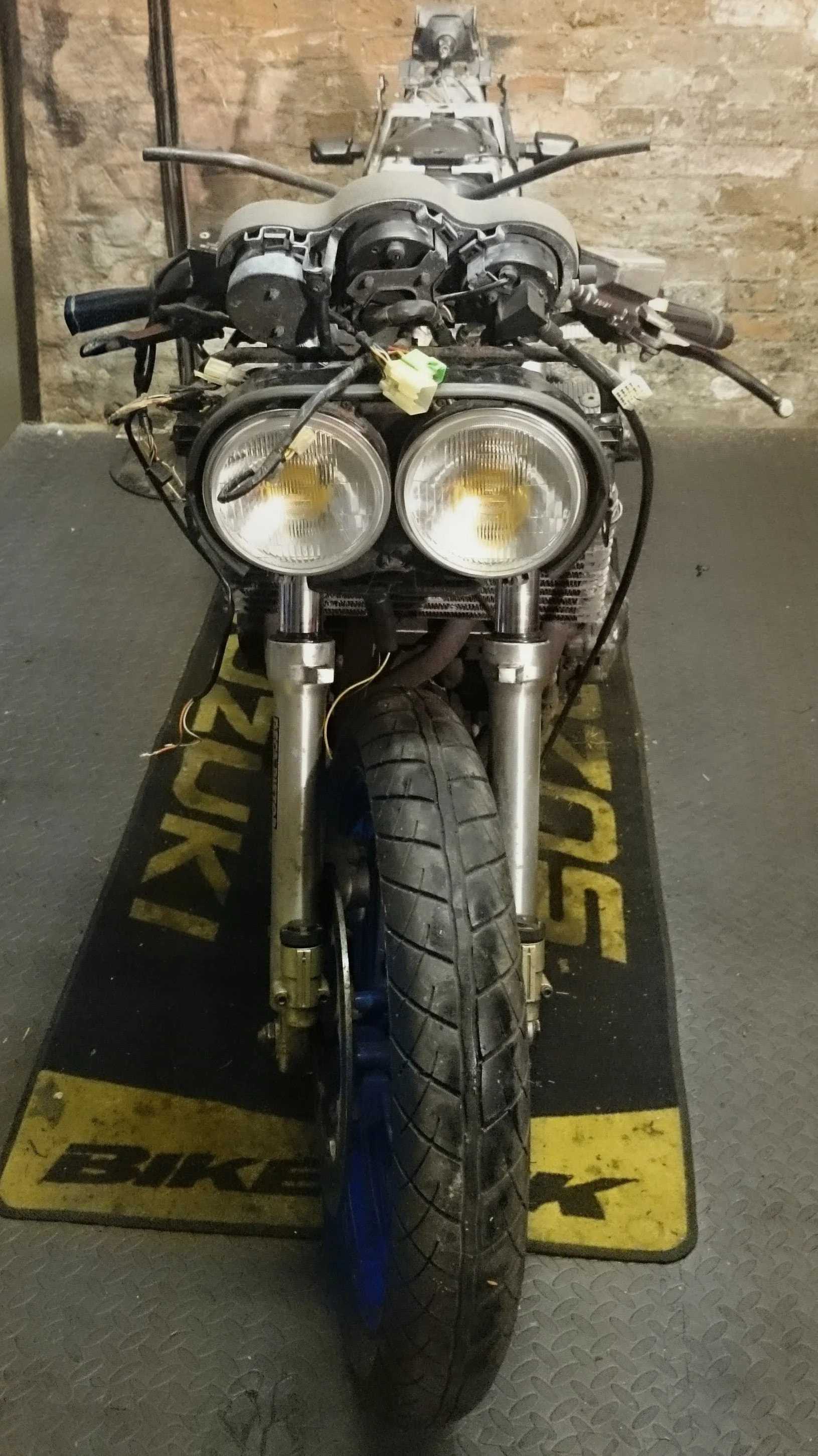 Front view of a partially disassembled motorcycle with exposed wiring and headlights, on a black and yellow mat in front of a brick wall.