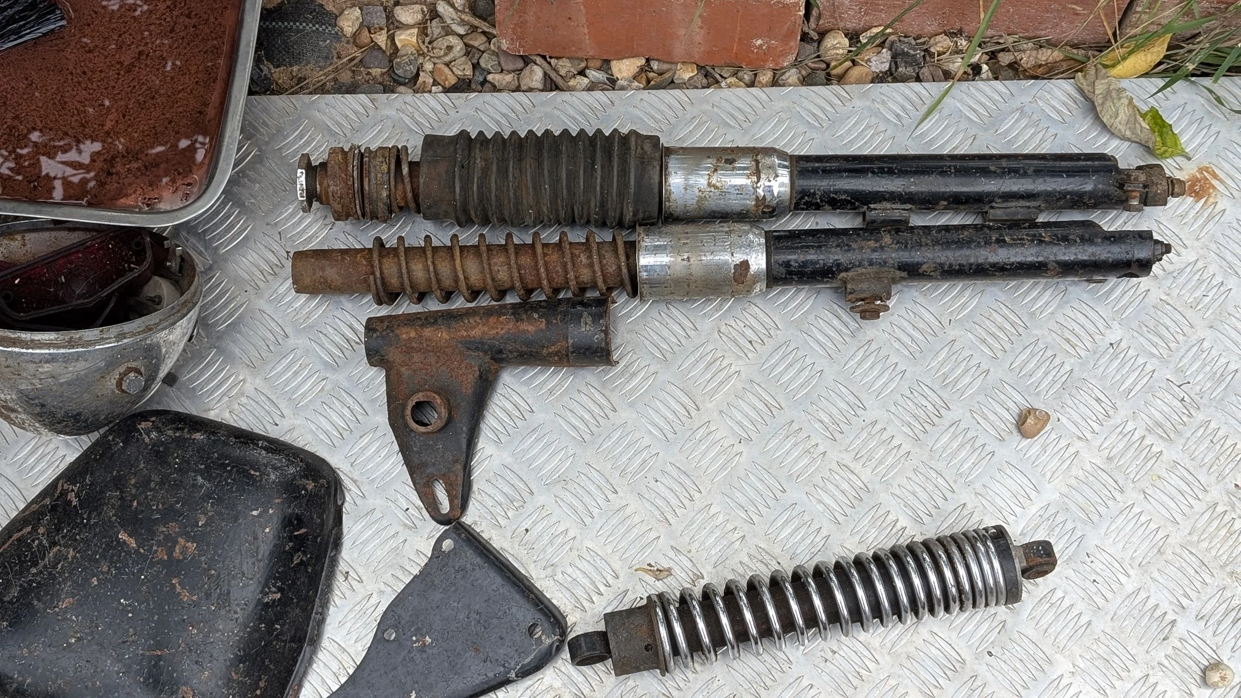 Various old, rusty and worn shock absorbers, forks and suspension parts laid out on a metal surface outdoors. Barn find Triumph T100 restoration project. 