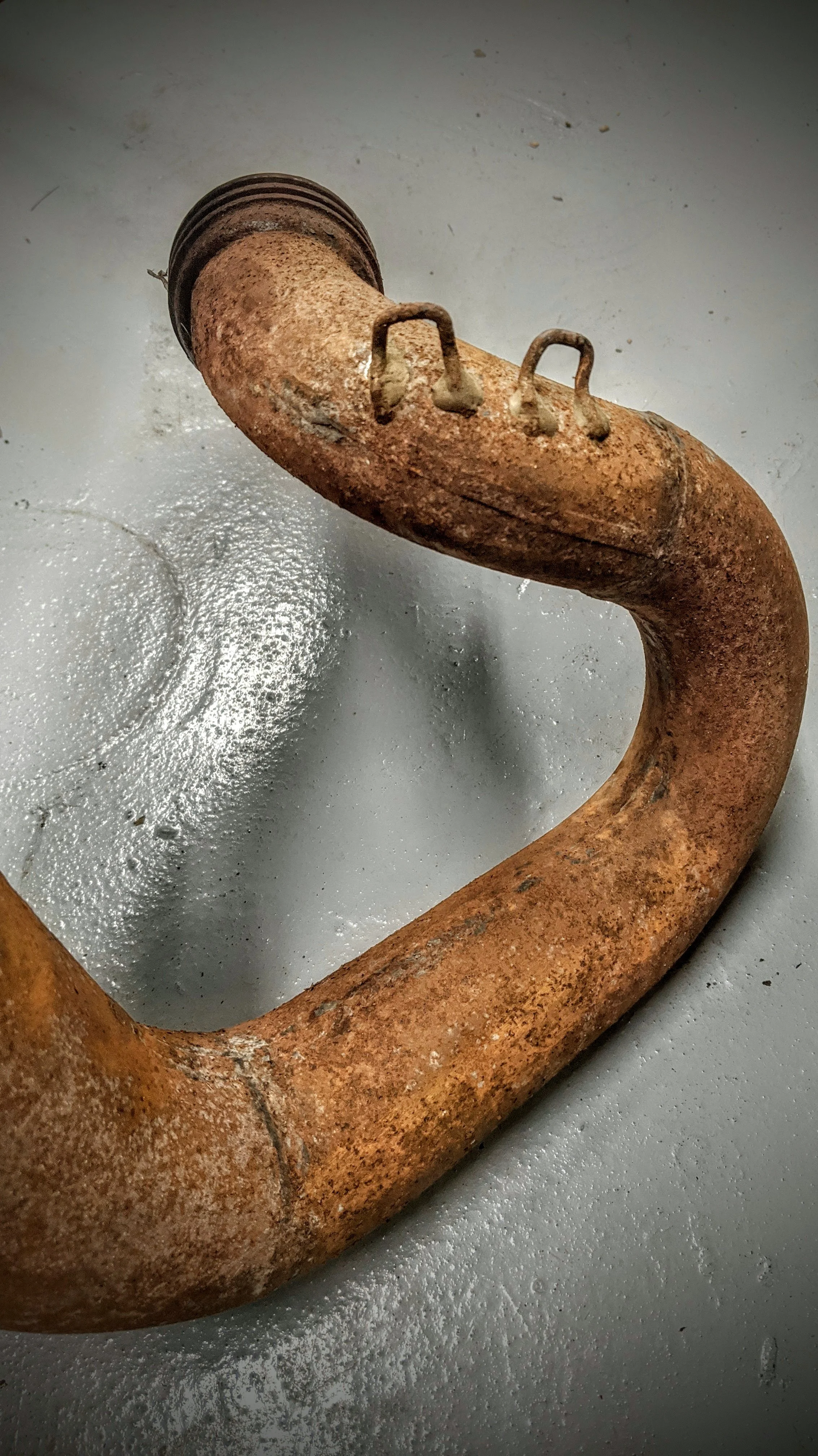 Close-up of a rusty, old car bumper with metal hooks, lying on a grey concrete floor with scattered white debris.