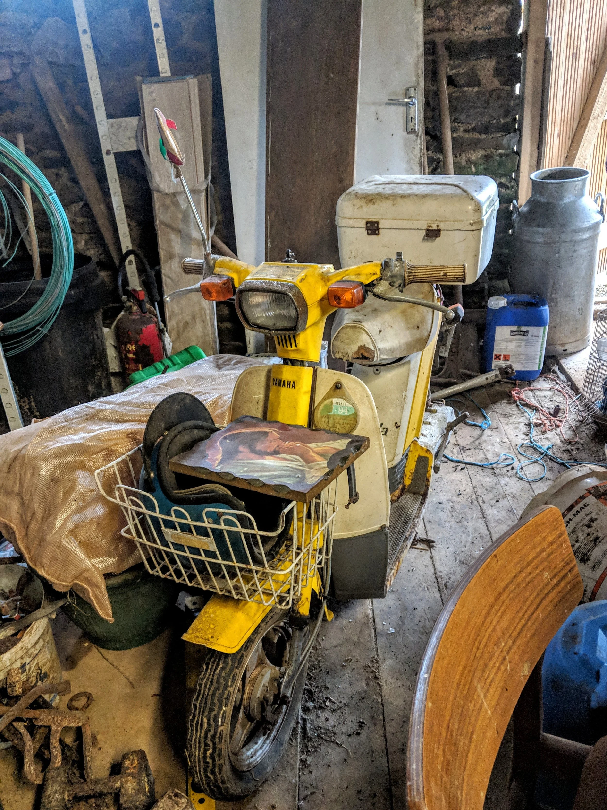 A vintage yellow Yamaha motor scooter with dents and rust parked inside a cluttered garage. The scooter has a white storage box attached to the back, and a small religious picture on the front. Surrounding it are various items, including a wooden chair, a black barrel, a blue container, and miscellaneous tools and wires, with a stone wall and wooden beams in the background.