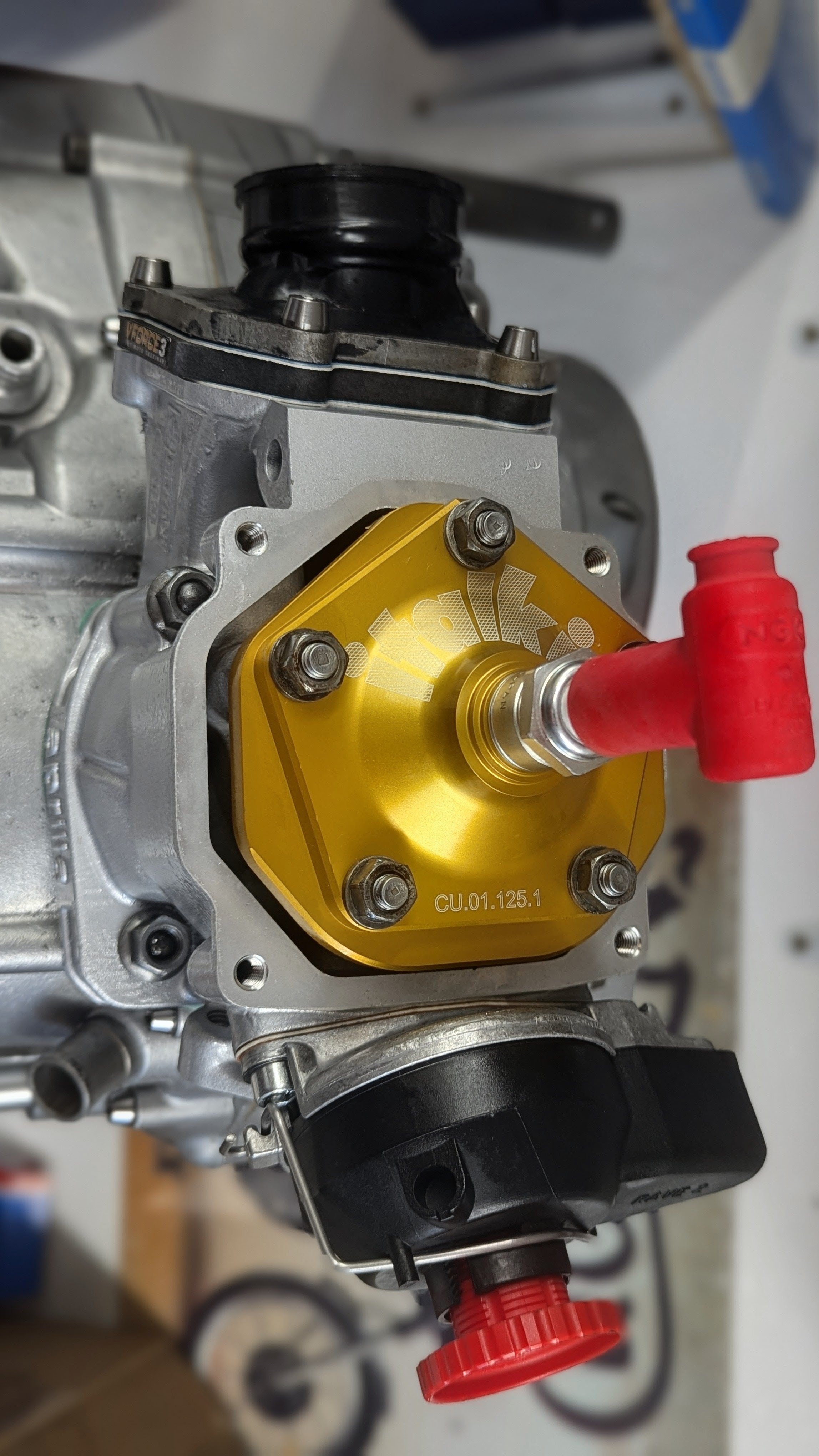 Close-up of a carburetor with a yellow K&N air filter adapter and red fuel line fittings.