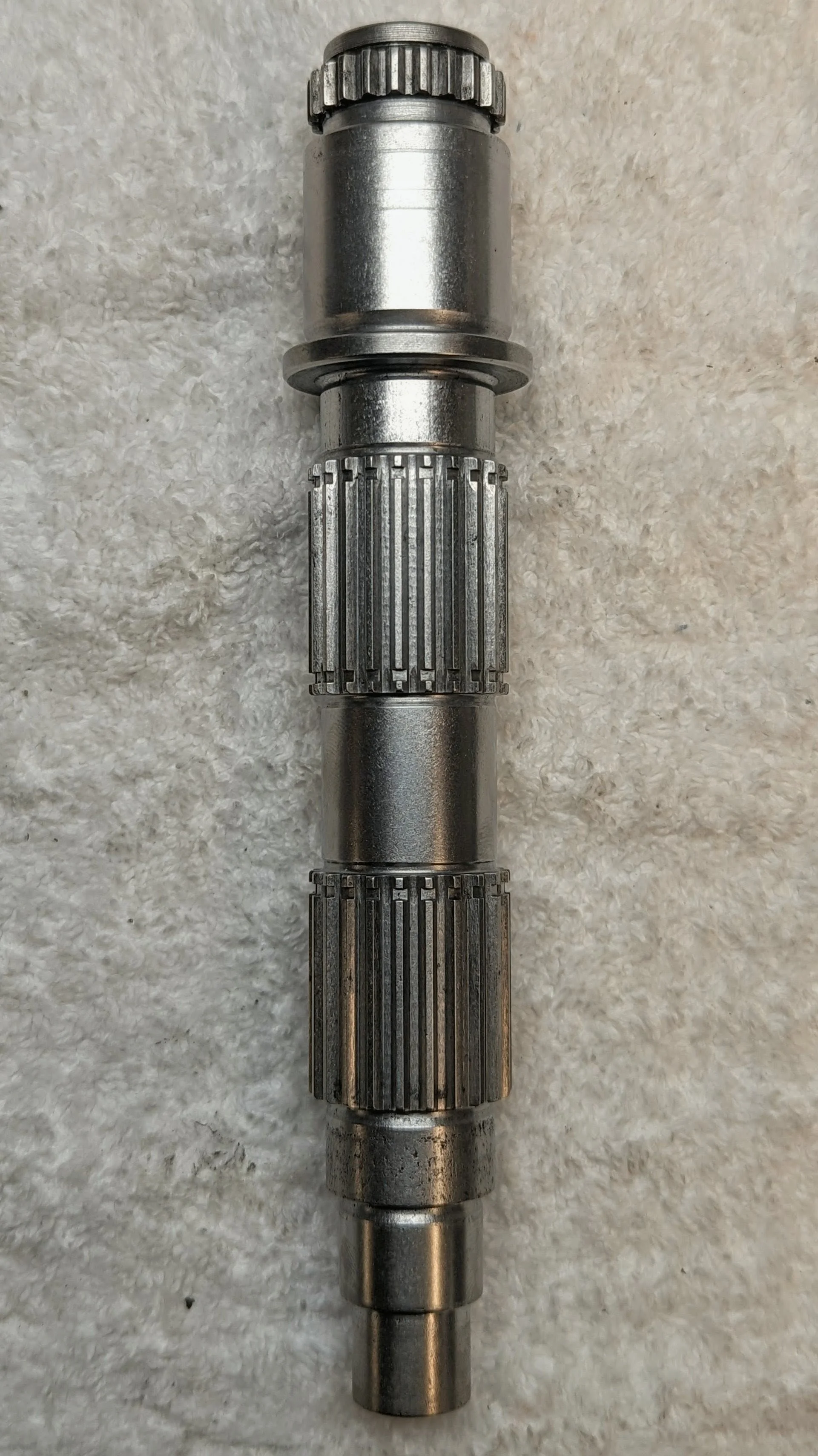 A metal gear shaft with splined ends lying on a white, textured surface.