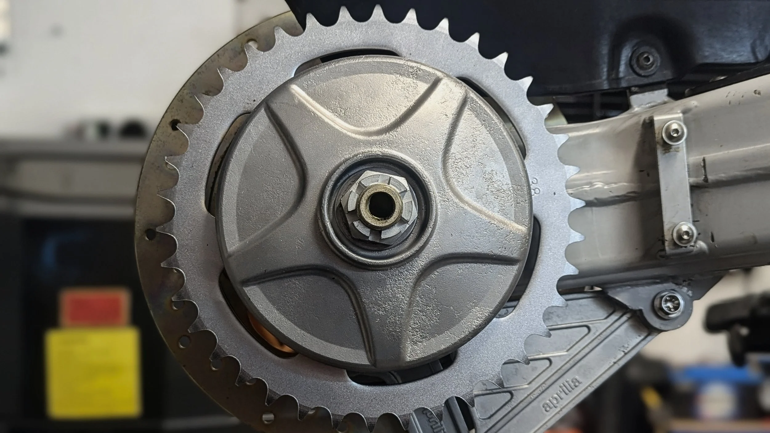 A close-up view of a motorcycle engine component, showing a large gear or sprocket with teeth, surrounded by metallic parts and fasteners.